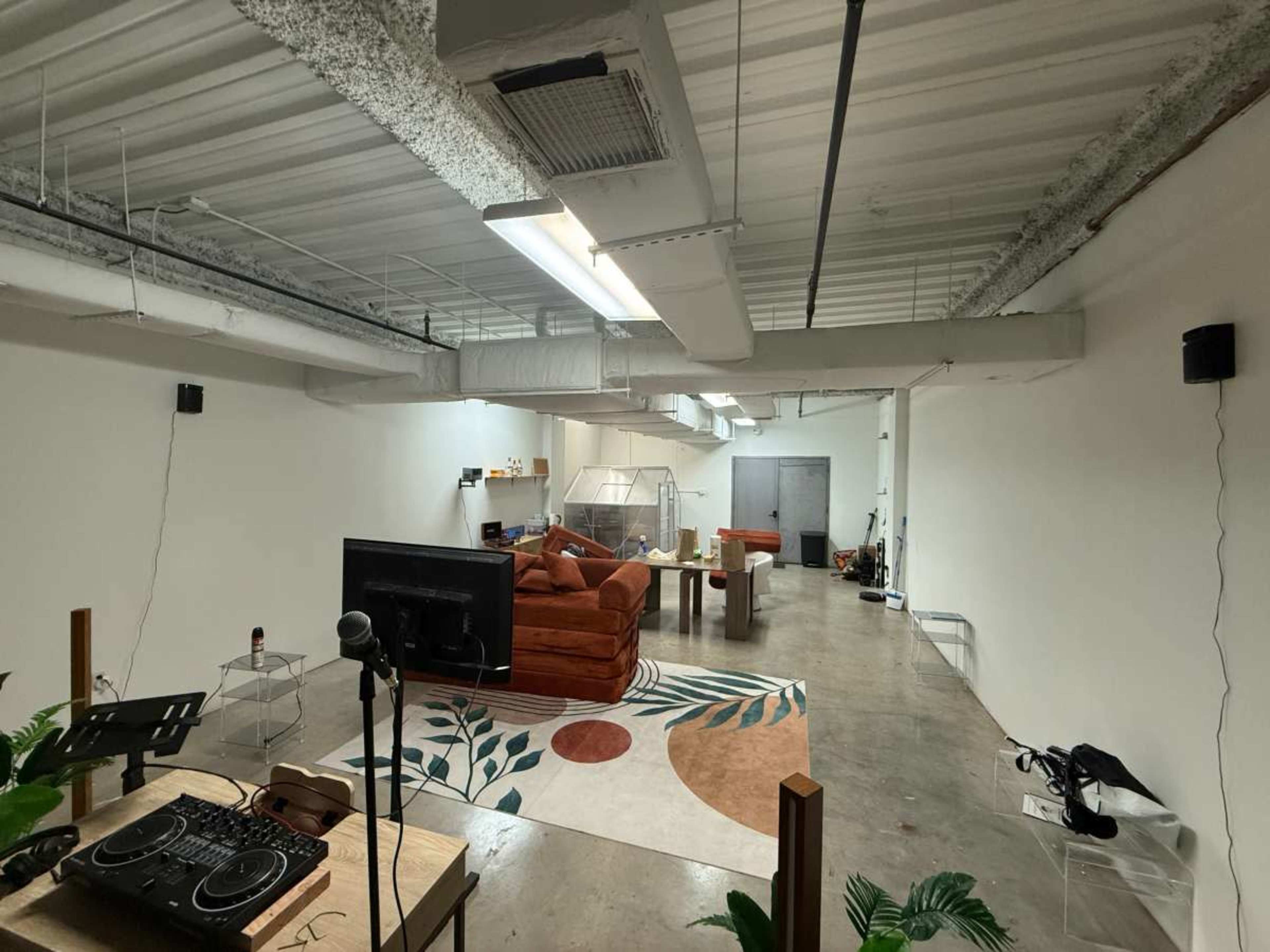 The image shows a spacious, minimally furnished room featuring a large orange couch, a coffee table with a microphone, and a patterned rug on a concrete floor.