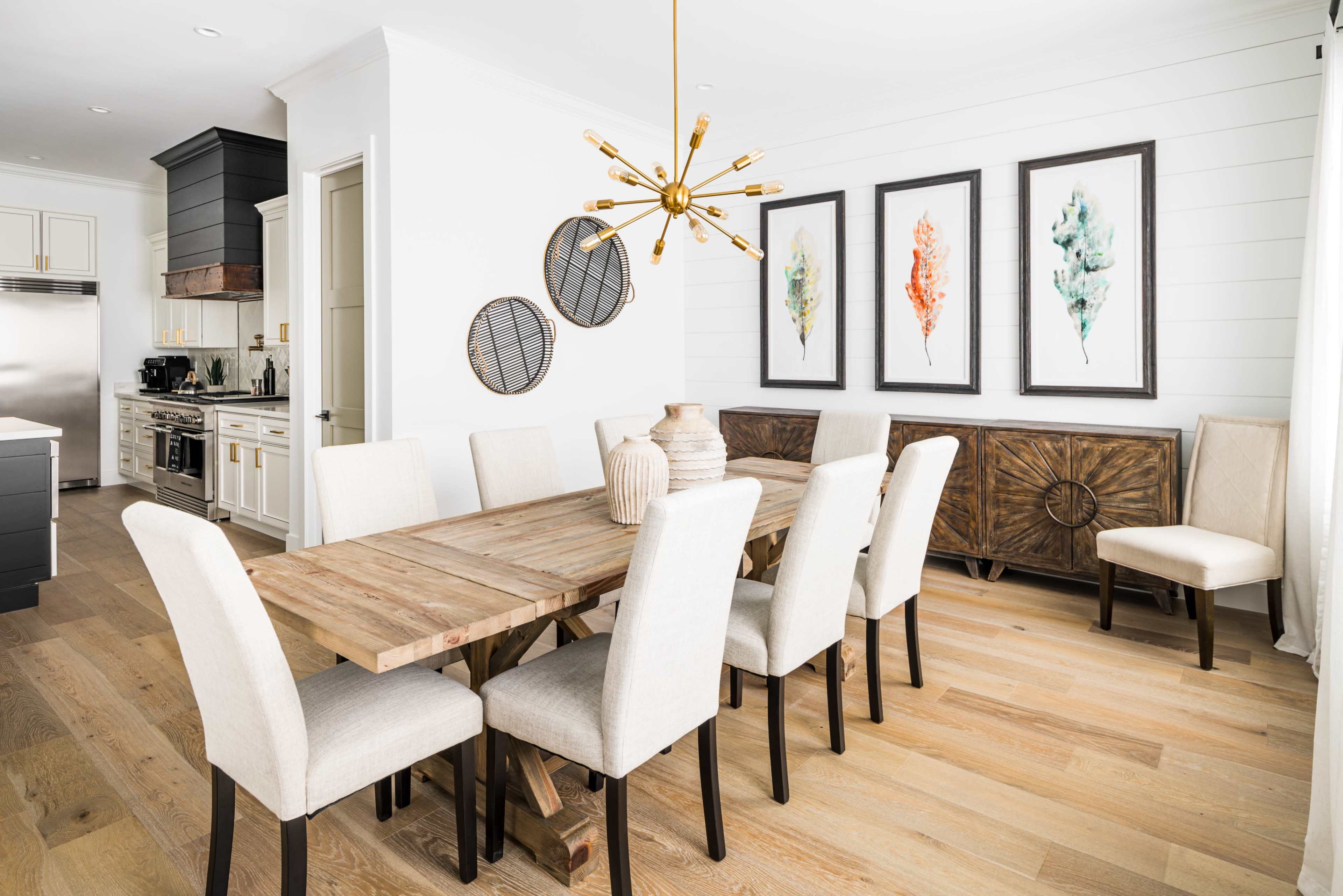 A wooden dining table with upholstered chairs is positioned in a bright room featuring framed artwork and a decorative sideboard against a light-colored wall.