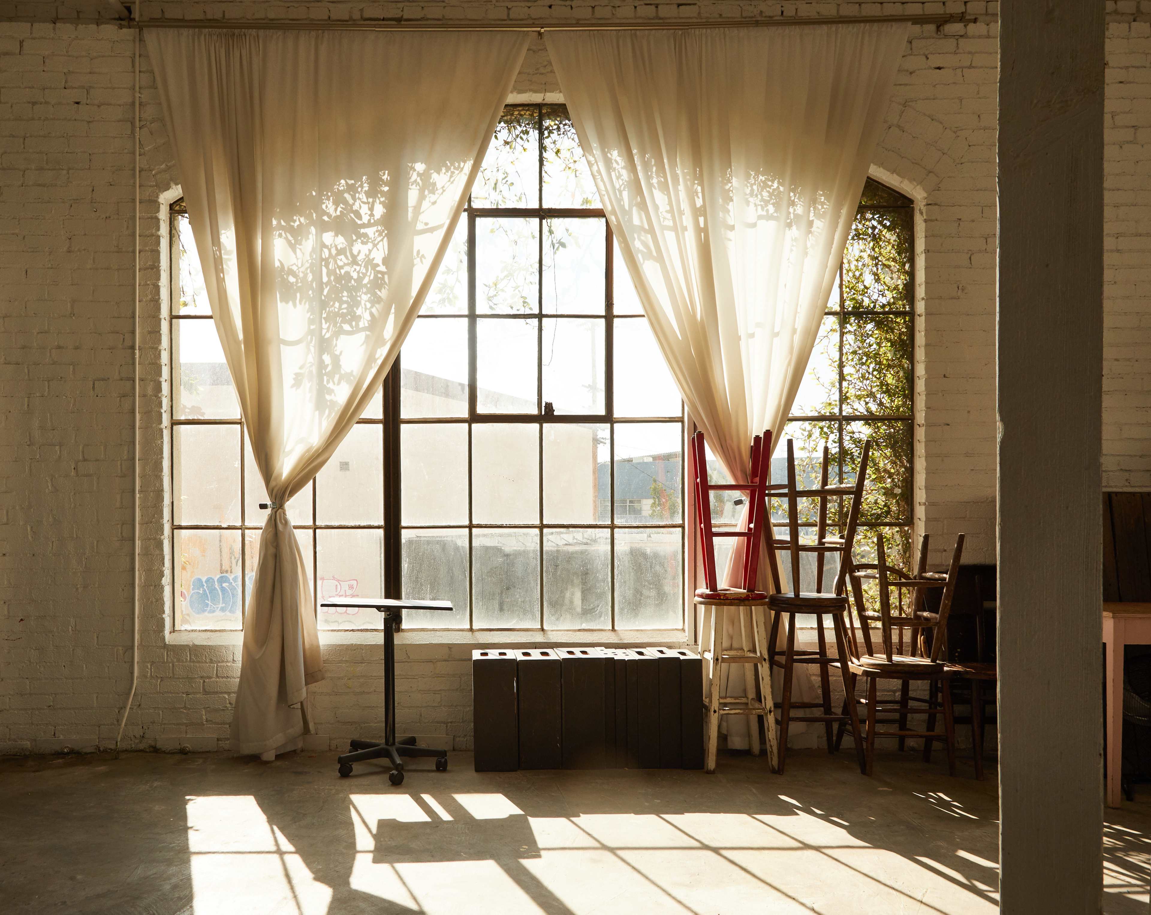 Sunlight filters through sheer curtains at a large window, illuminating a room furnished with various chairs and a small table.