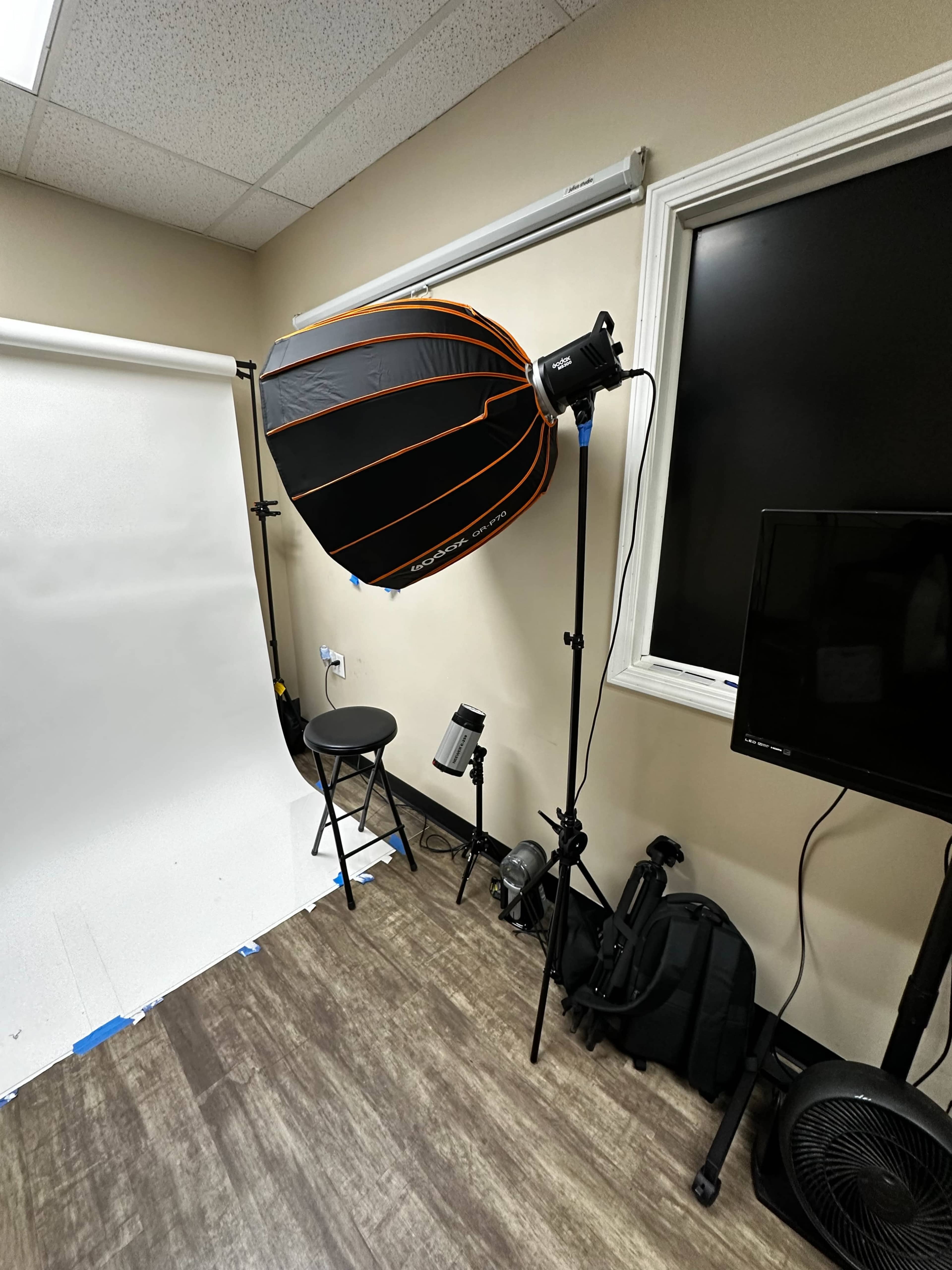 A photography studio setup featuring a large softbox light, a stool, a background roll, and various photography equipment along the walls.