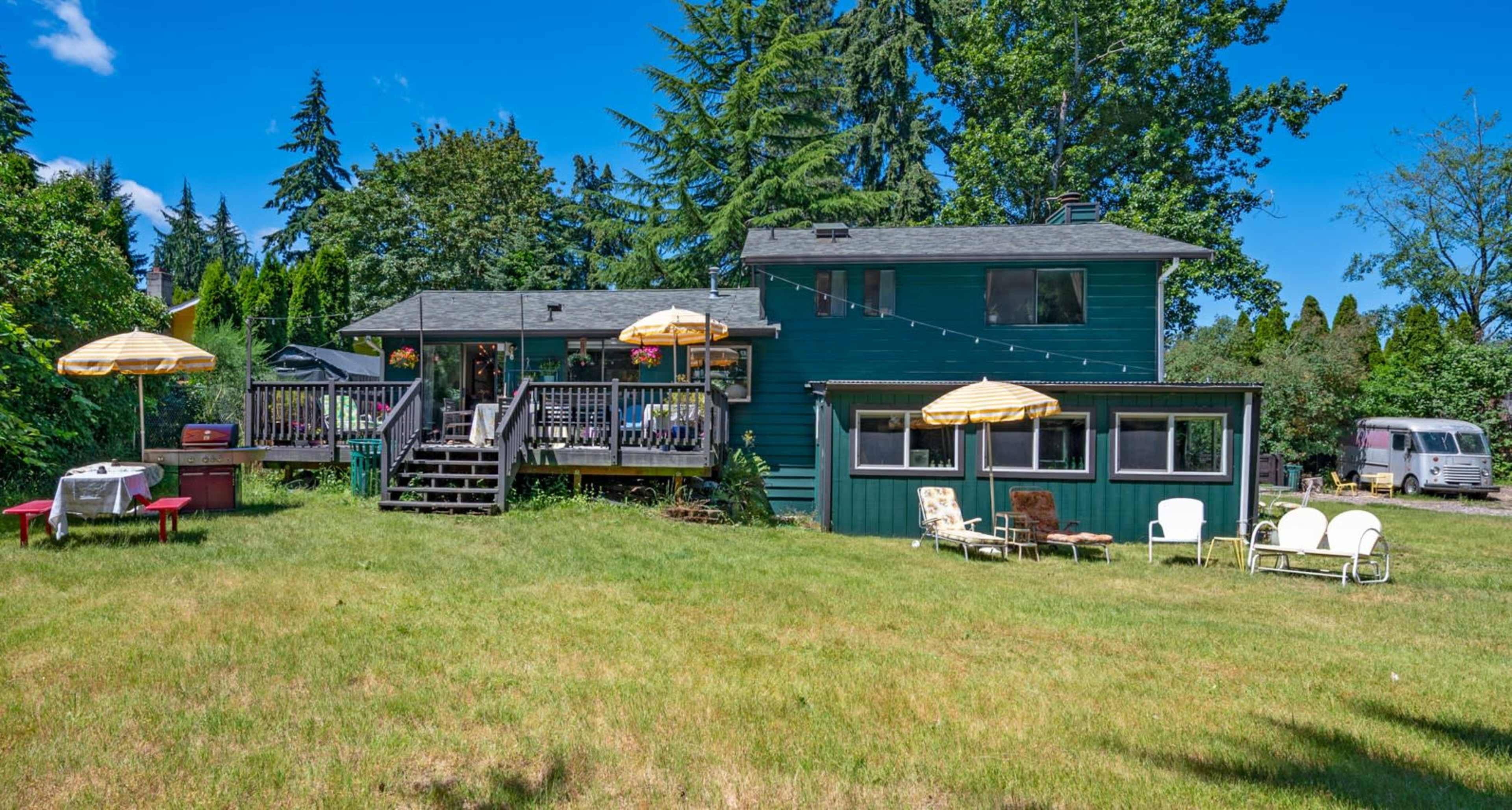 A two-story green house with a deck and several outdoor seating areas is set in a grassy yard surrounded by trees.