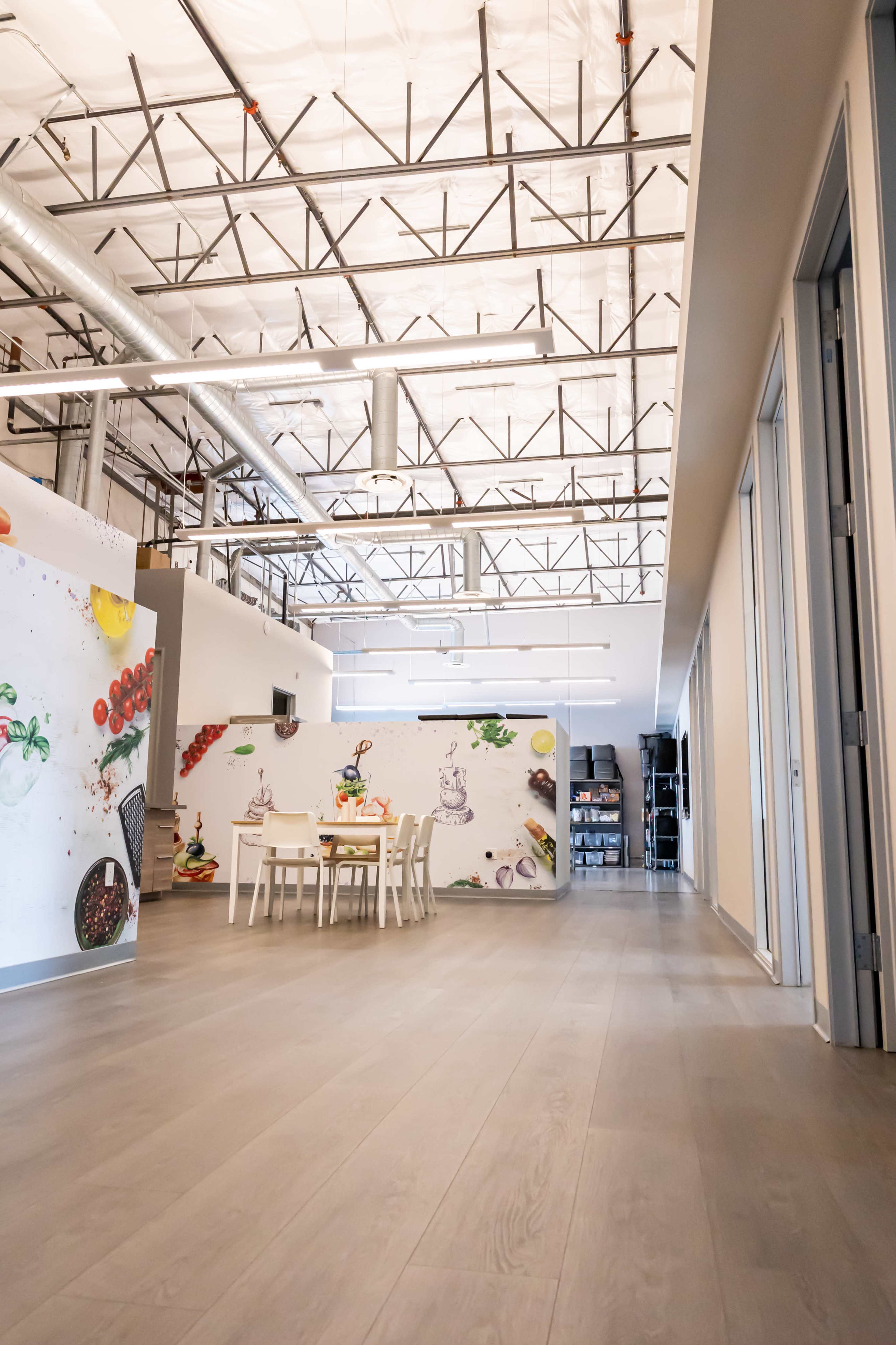 The image shows a spacious, modern office hallway with a dining area featuring a table and chairs, and a wall decorated with colorful illustrations of food items.