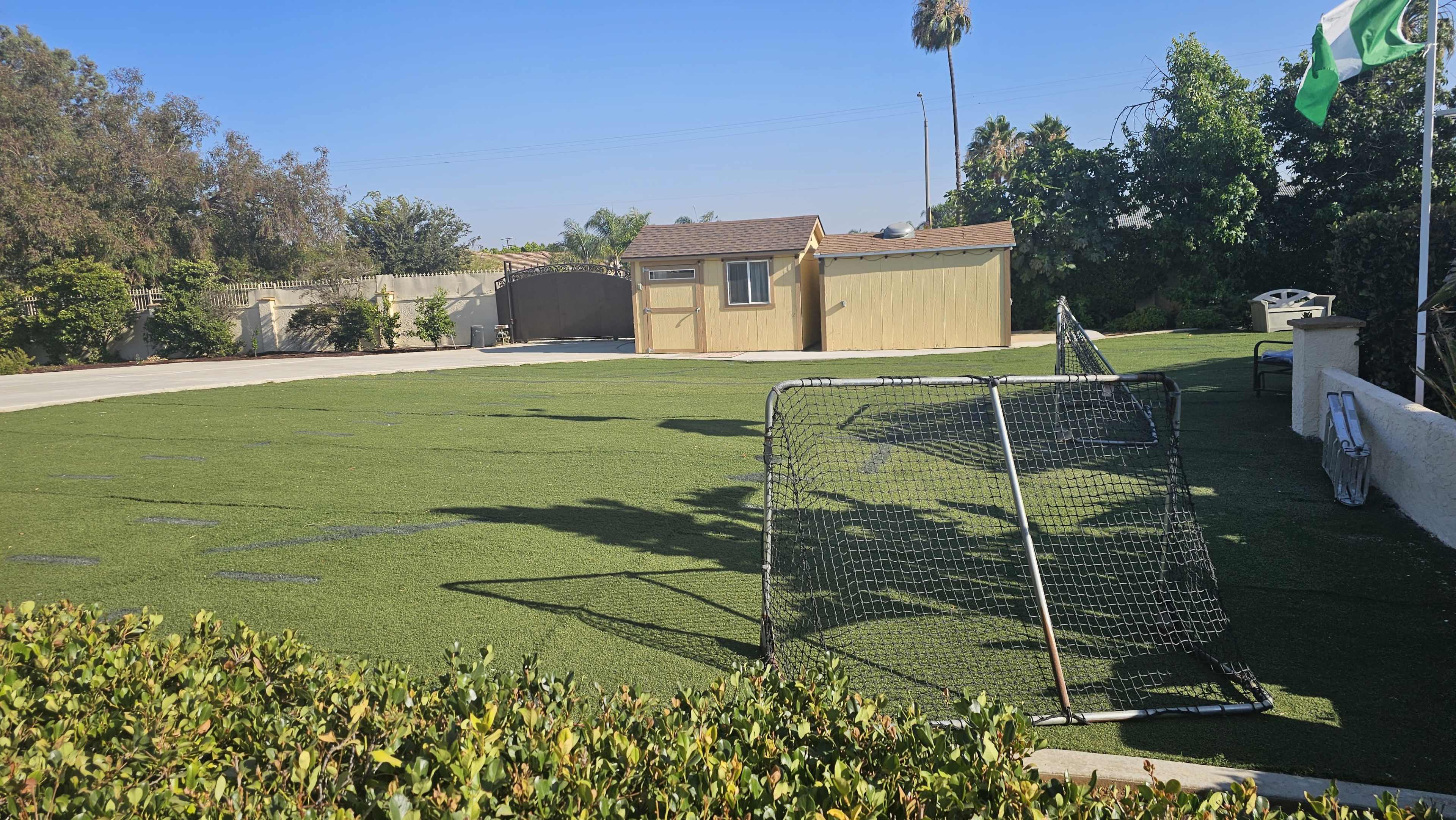 The image shows a green artificial turf area with two soccer goals, a small yellow shed in the background, and a fence with flags.