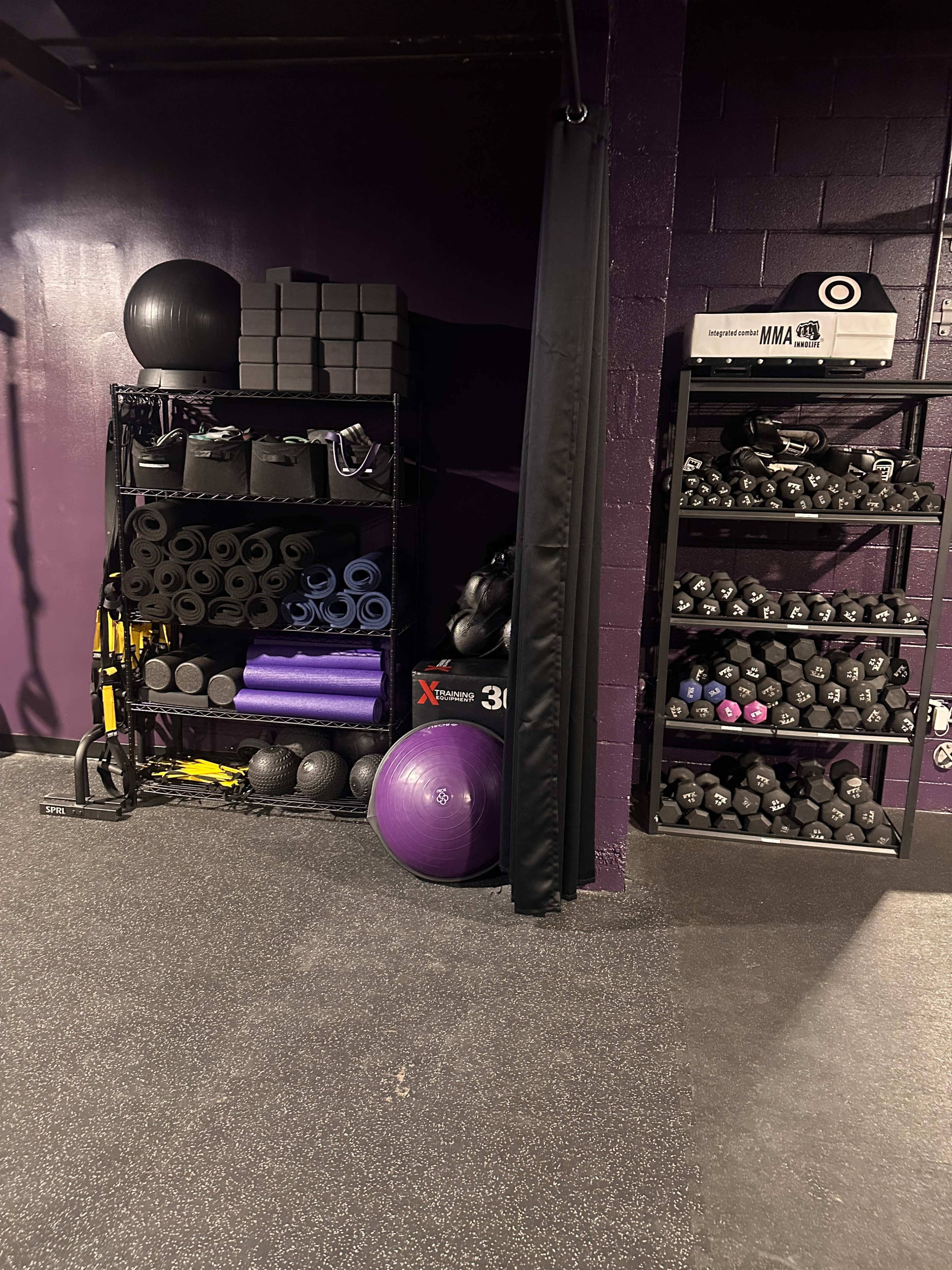 The image shows a gym storage area with shelves filled with exercise equipment such as stability balls, foam rollers, weights, and resistance bands, against a purple wall.