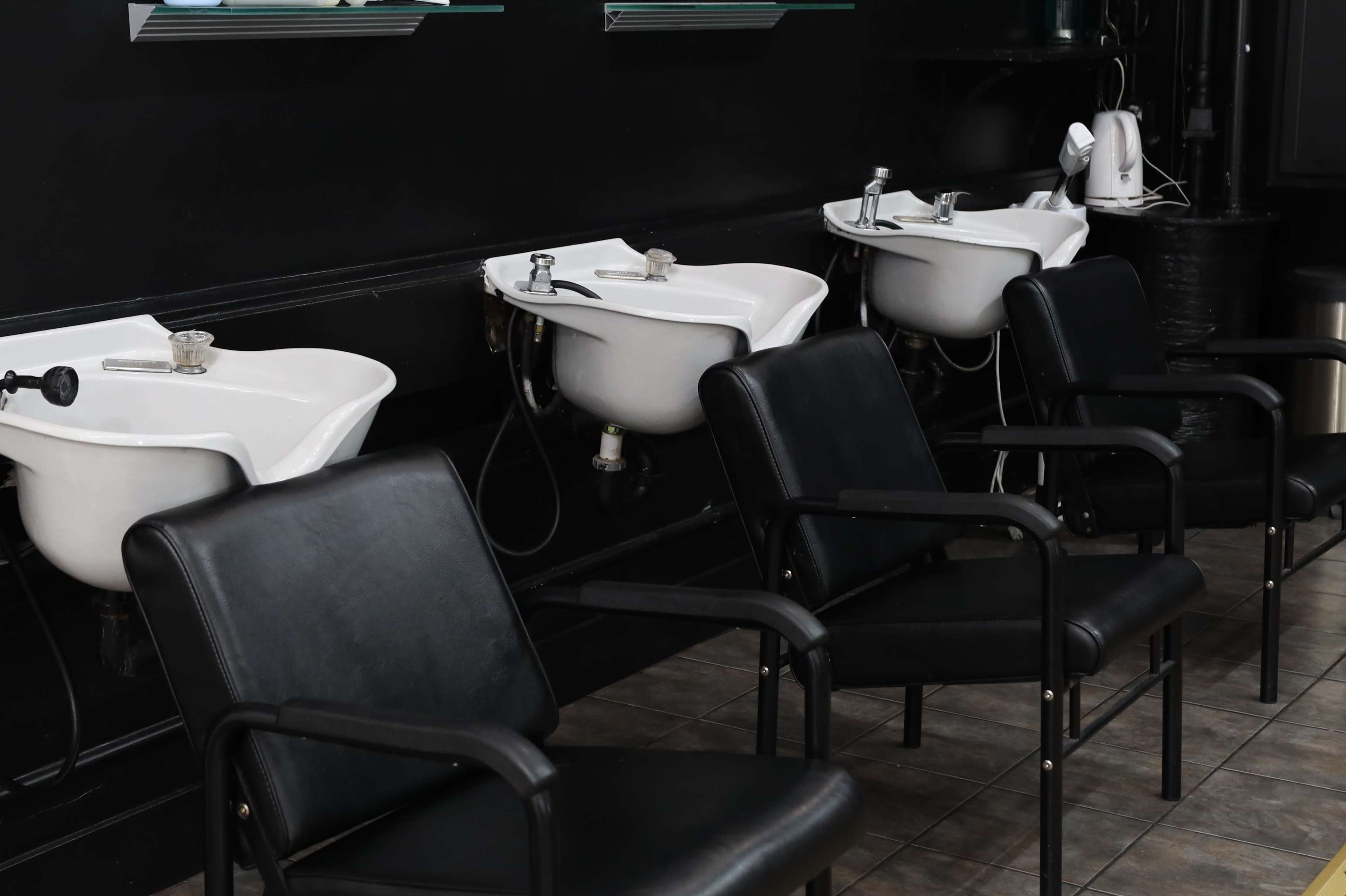 Three white shampoo bowls lined against a black wall, accompanied by black chairs in front of each bowl.