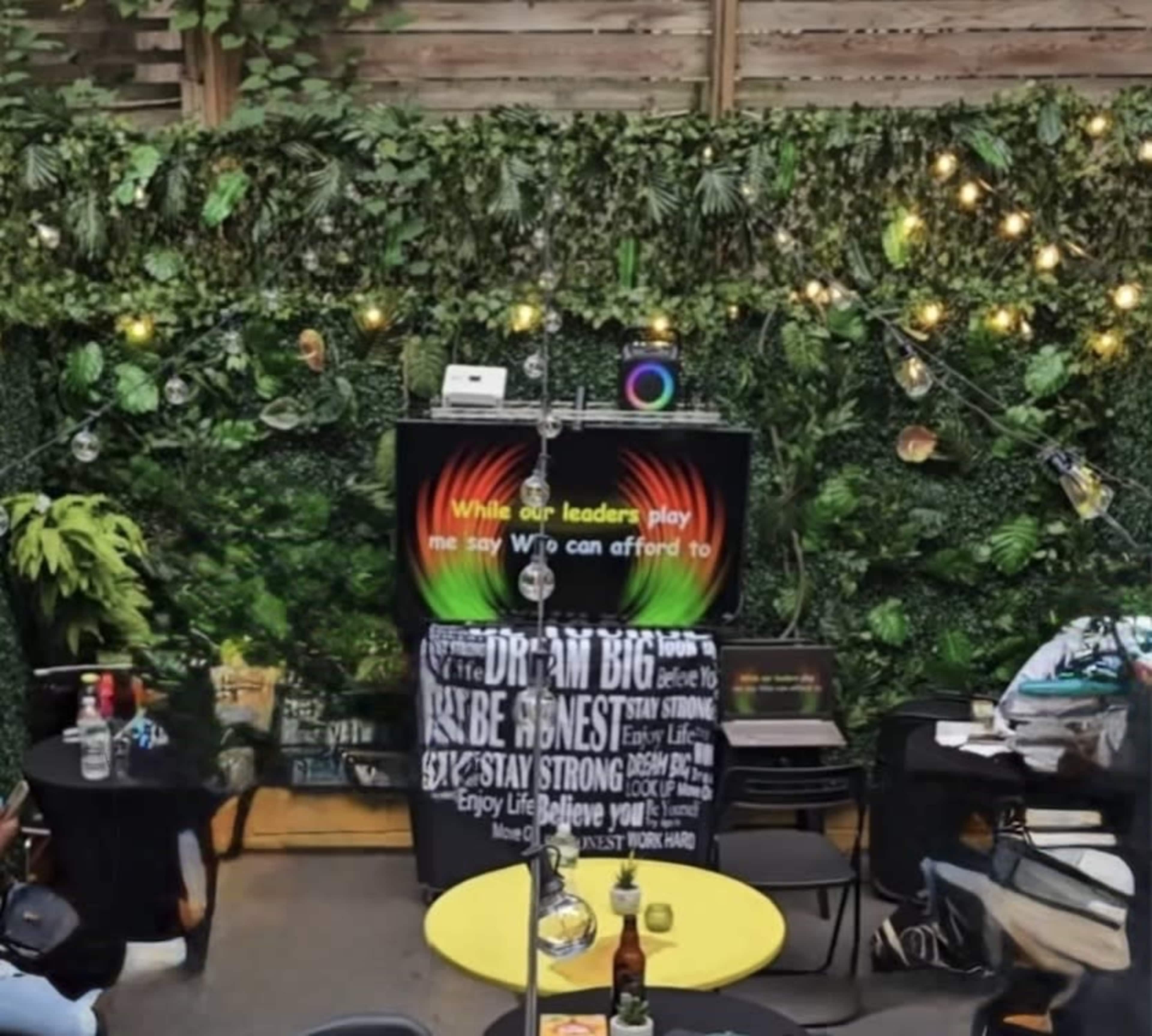 The image shows an outdoor space decorated with greenery and lights, featuring a stage setup with a speaker and a colorful display in the background.