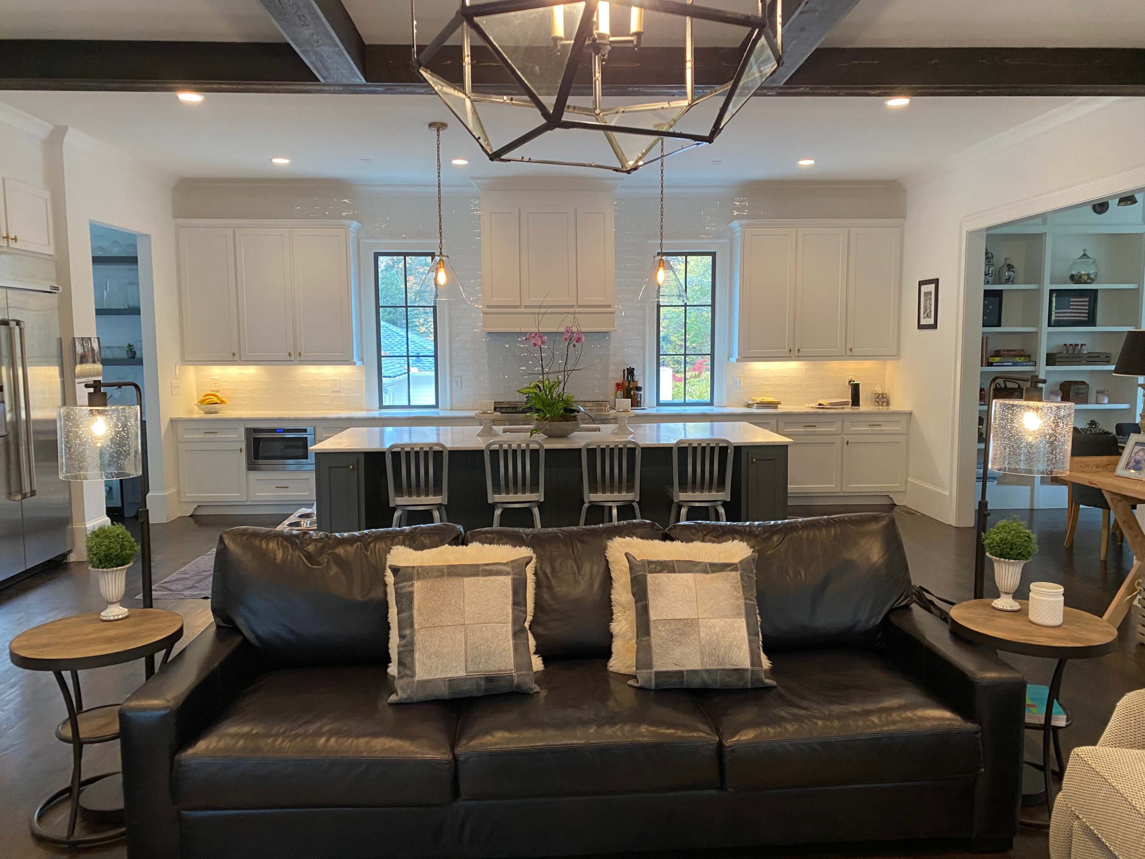 A black leather sofa with decorative pillows faces a modern kitchen featuring white cabinets and a large island with bar stools.