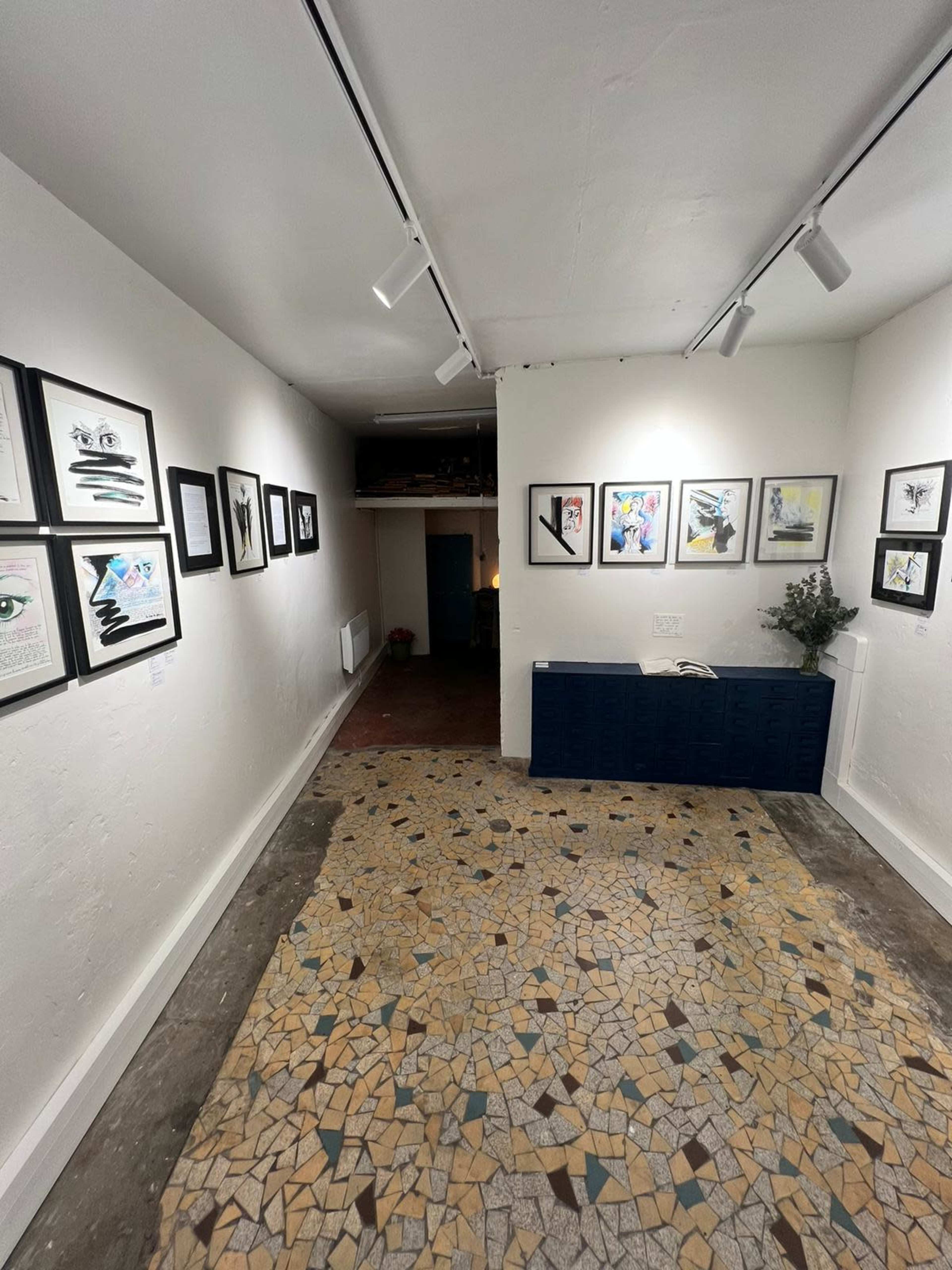 The image shows a narrow gallery space with art displayed on the walls and a patterned floor.