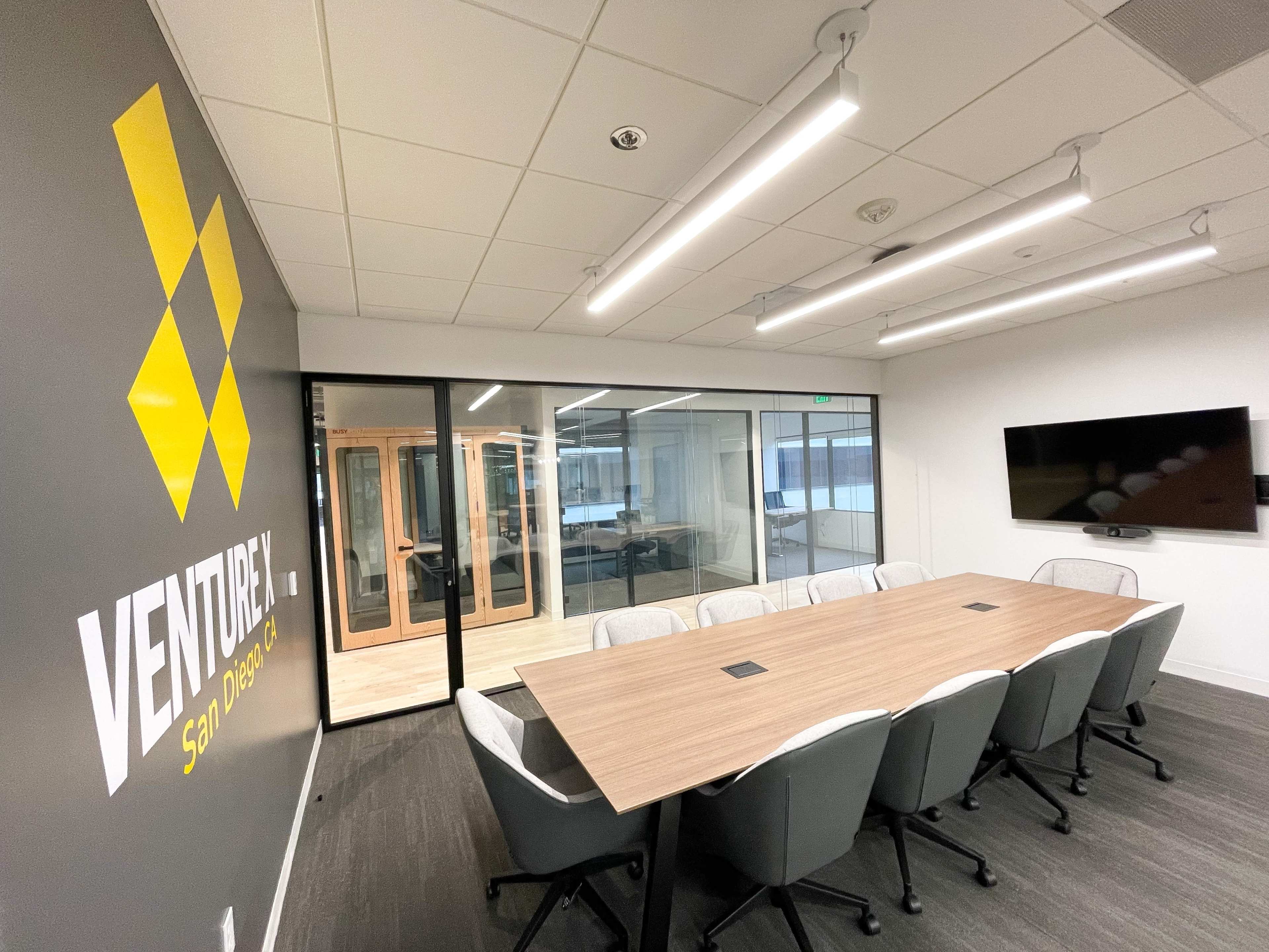 A spacious conference room features a long wooden table, ergonomic chairs, and glass walls with a logo displayed on the adjacent wall.