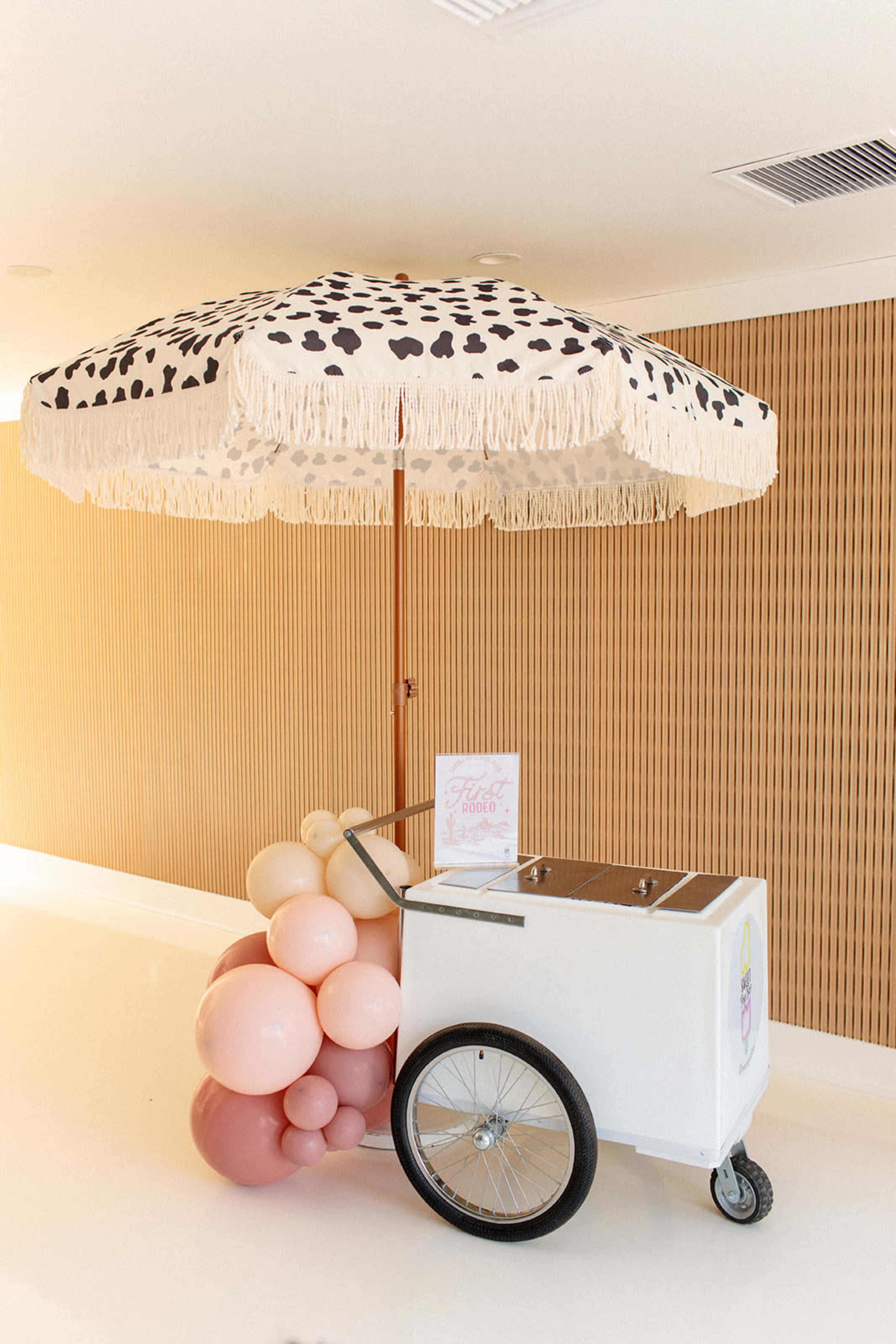 A cart with a vintage design is decorated with a large umbrella and is surrounded by clusters of balloons in shades of pink.