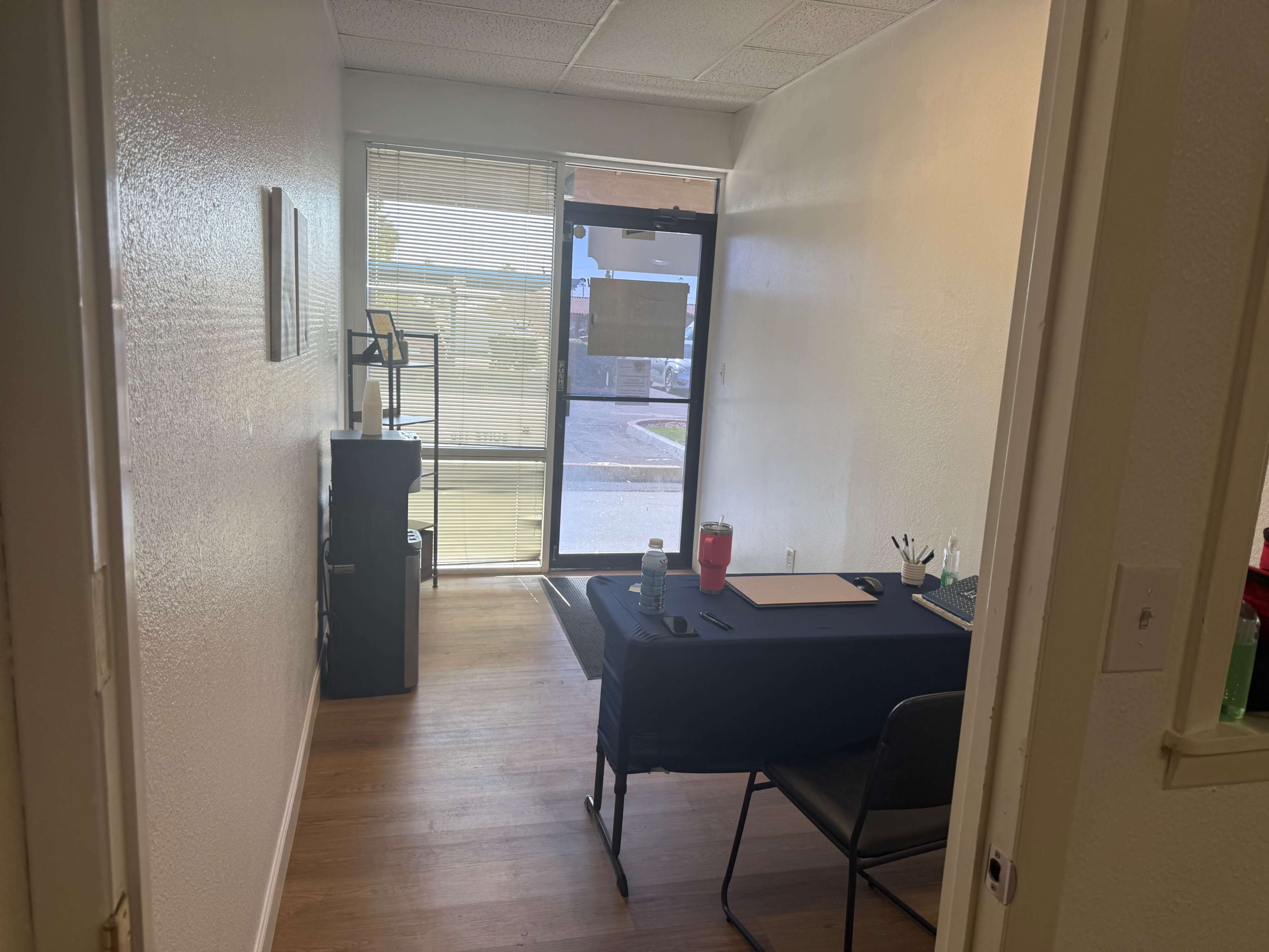 The image shows a small, well-lit office space with a black table, two chairs, and a door leading to an exterior area.