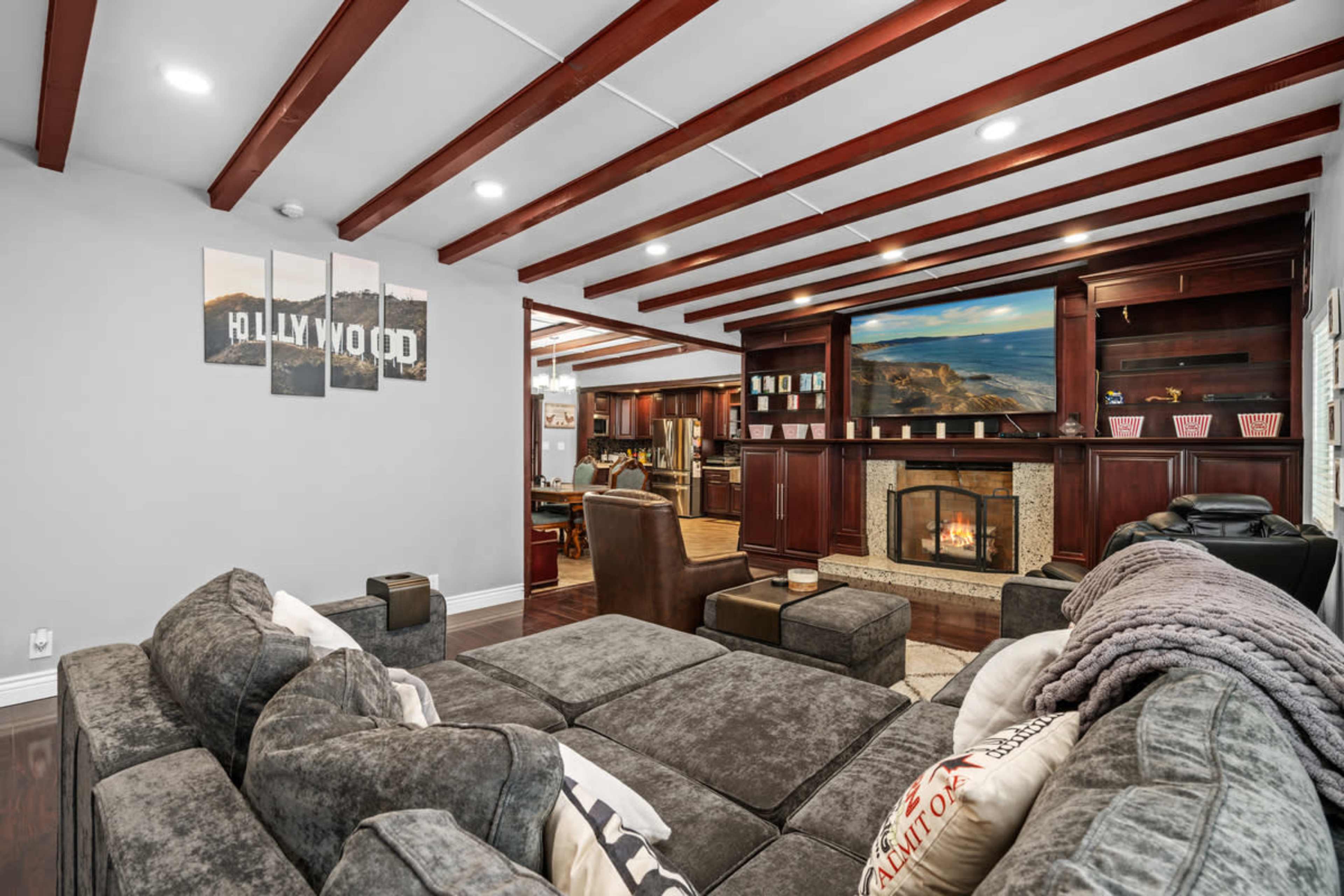 A modern living room with a stone fireplace, a sectional sofa, and beams on the ceiling, leading to a kitchen in the background.