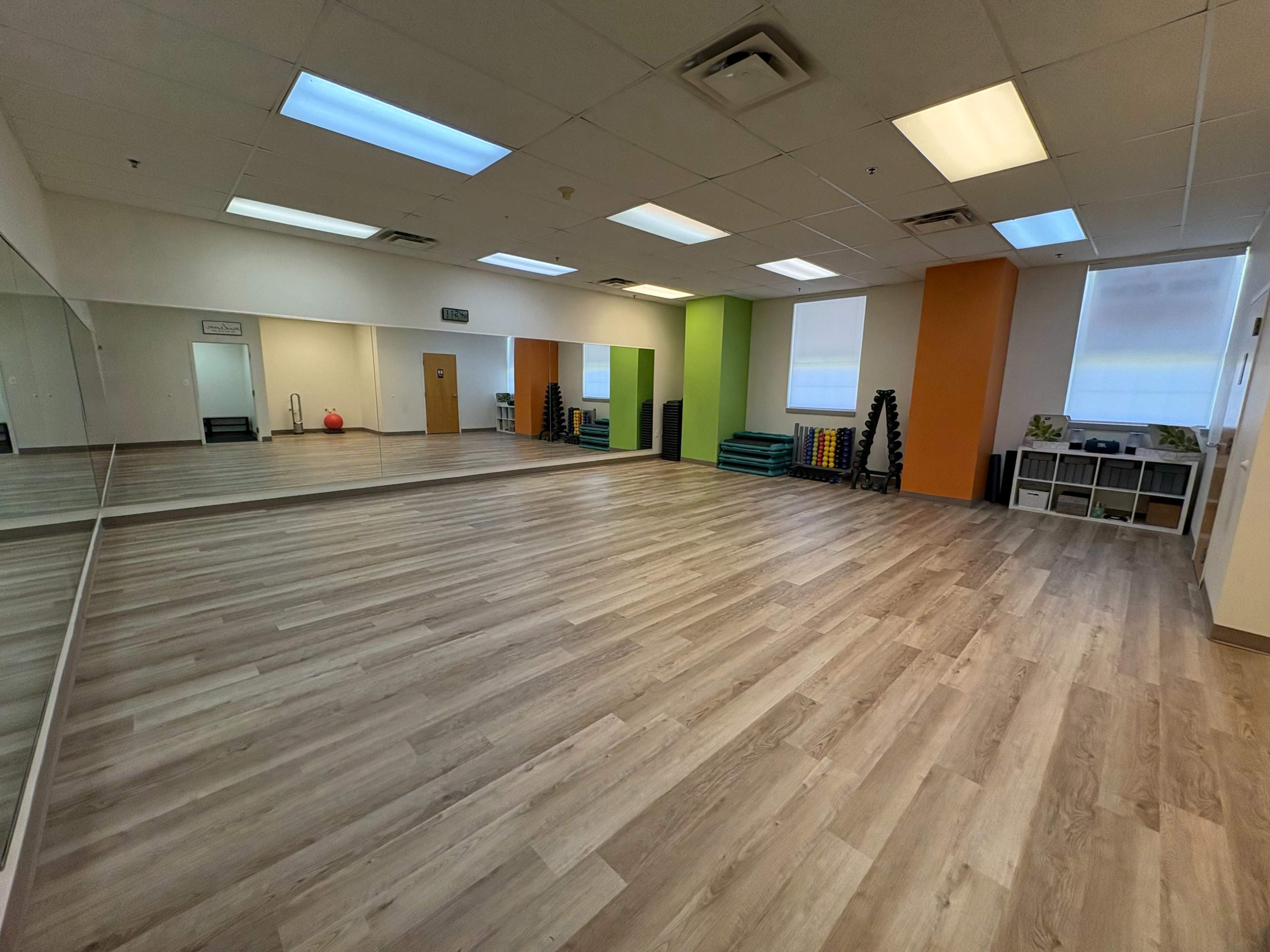 The image shows a spacious and well-lit exercise studio featuring wooden flooring, large mirrors, and colored walls with fitness equipment arranged along the sides.