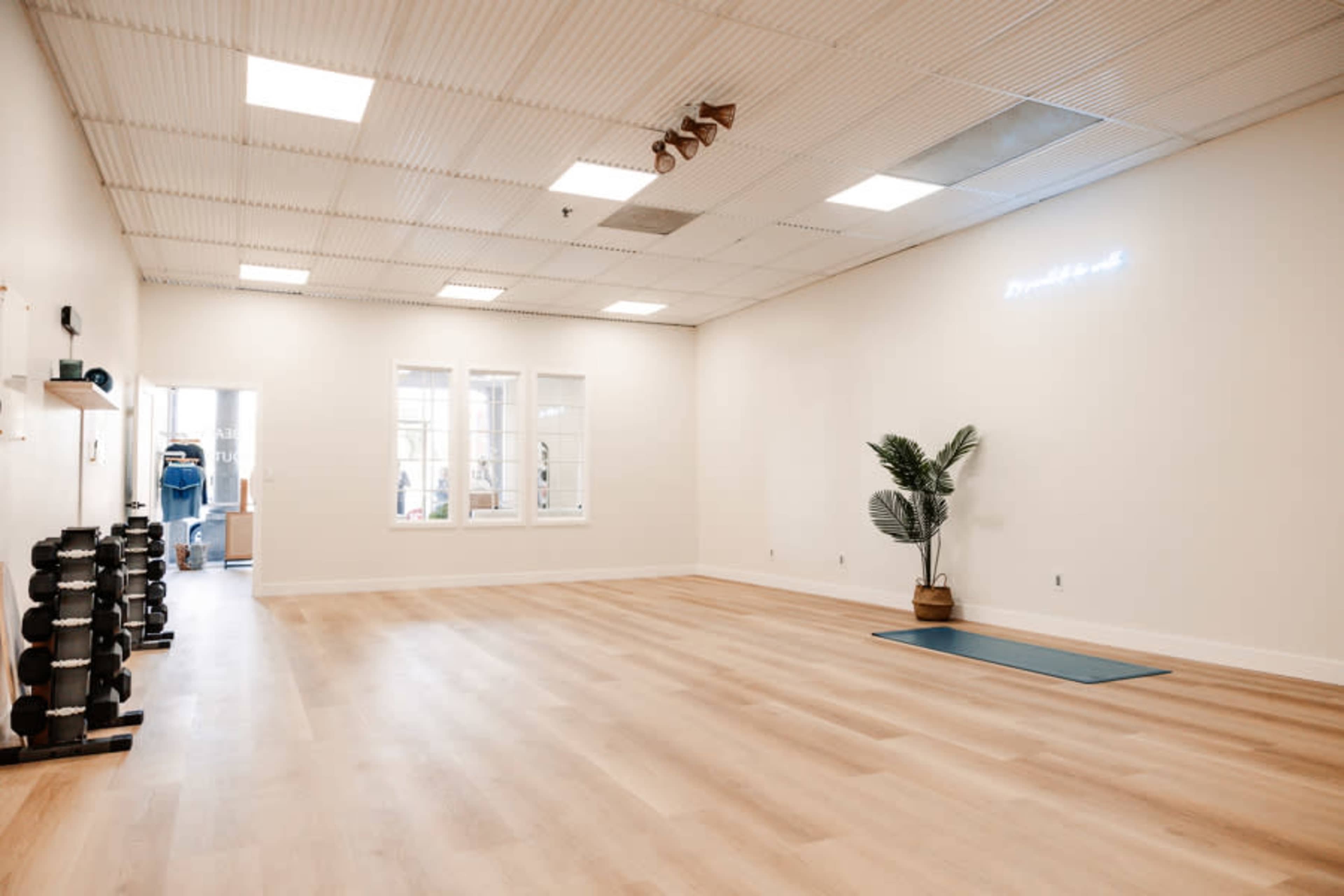 The image shows a spacious, well-lit fitness studio with wooden flooring, a yoga mat, a potted plant, and a set of dumbbells on a rack.