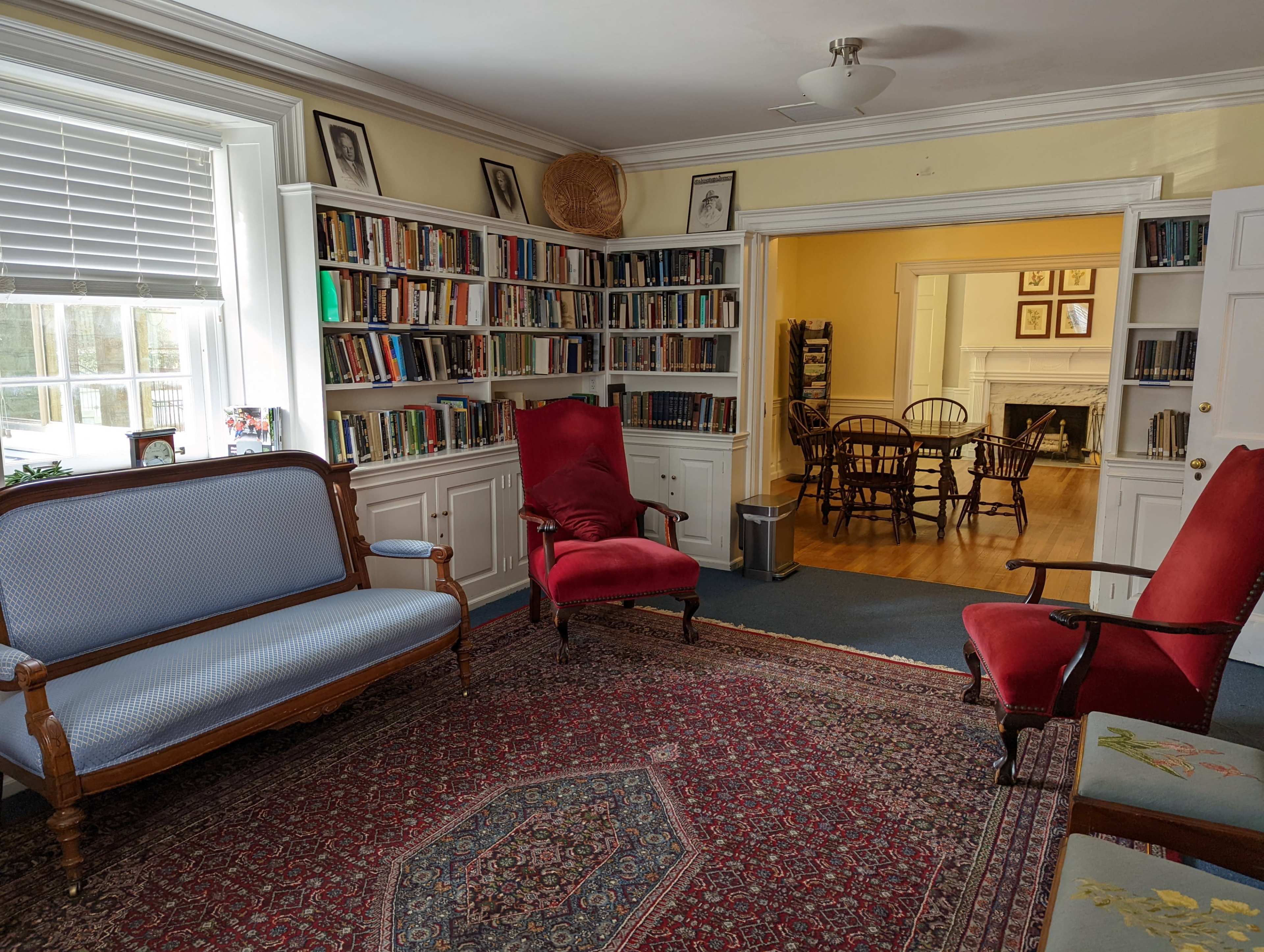 A cozy room with bookshelves lining the walls, two red chairs, a blue upholstered sofa, and a patterned rug, leading to a dining area with a wooden table and chairs visible in the background.