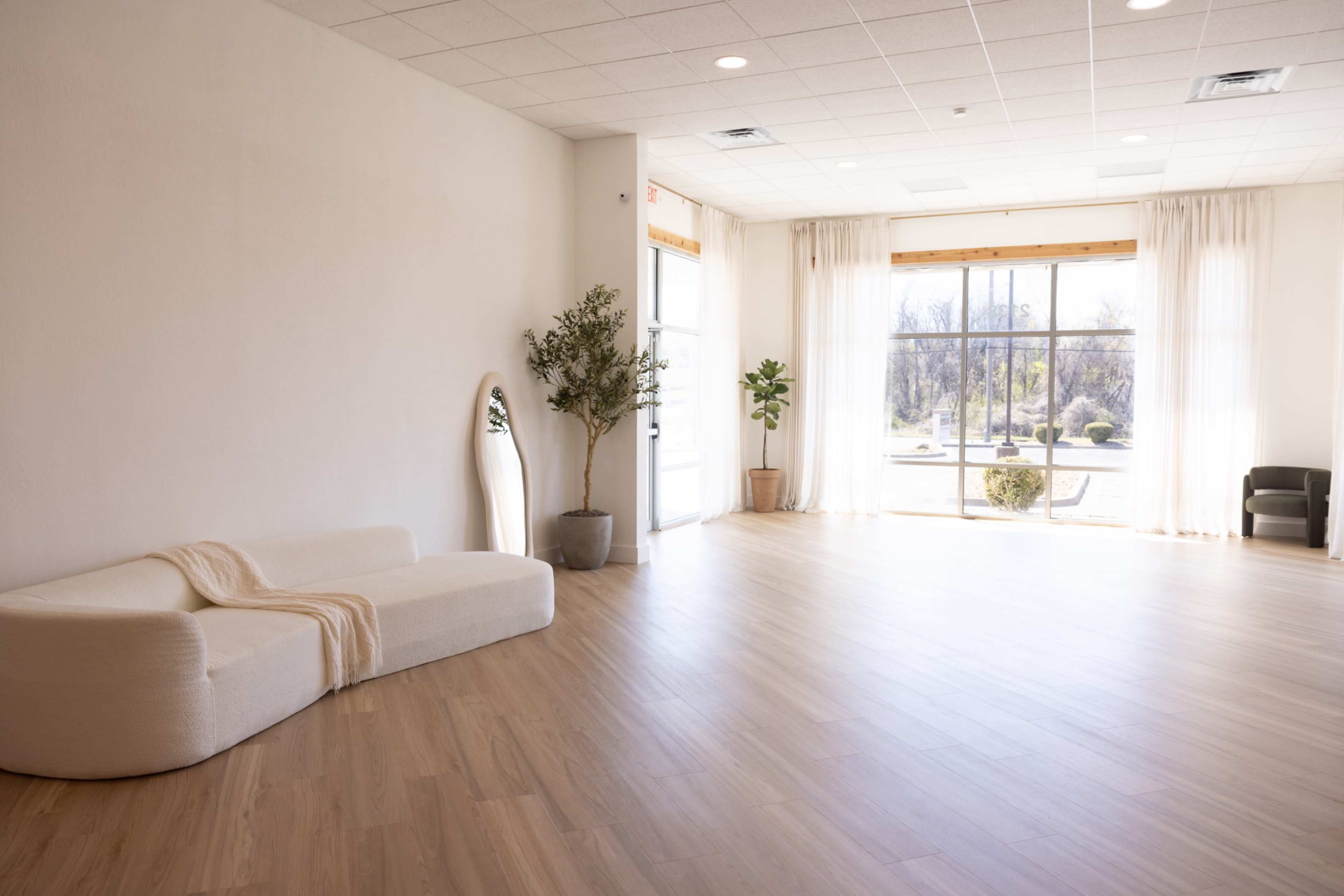 The image shows a spacious, well-lit room with light wooden flooring, minimal furniture, and large windows that provide a view of the outdoors.