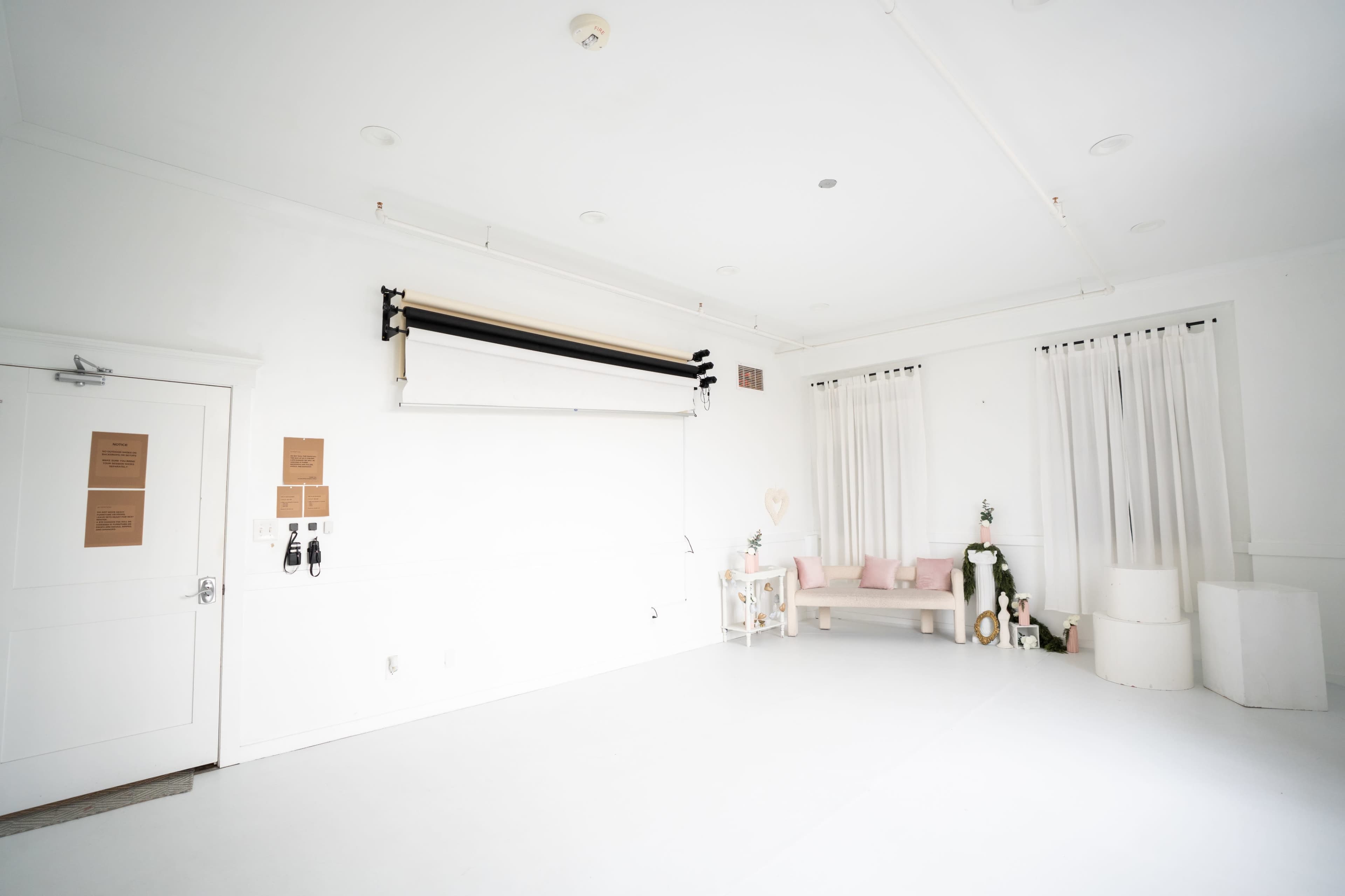 The image depicts a bright, minimalist studio space with white walls and flooring, featuring a backdrop support, a small seating area, and decorative elements.