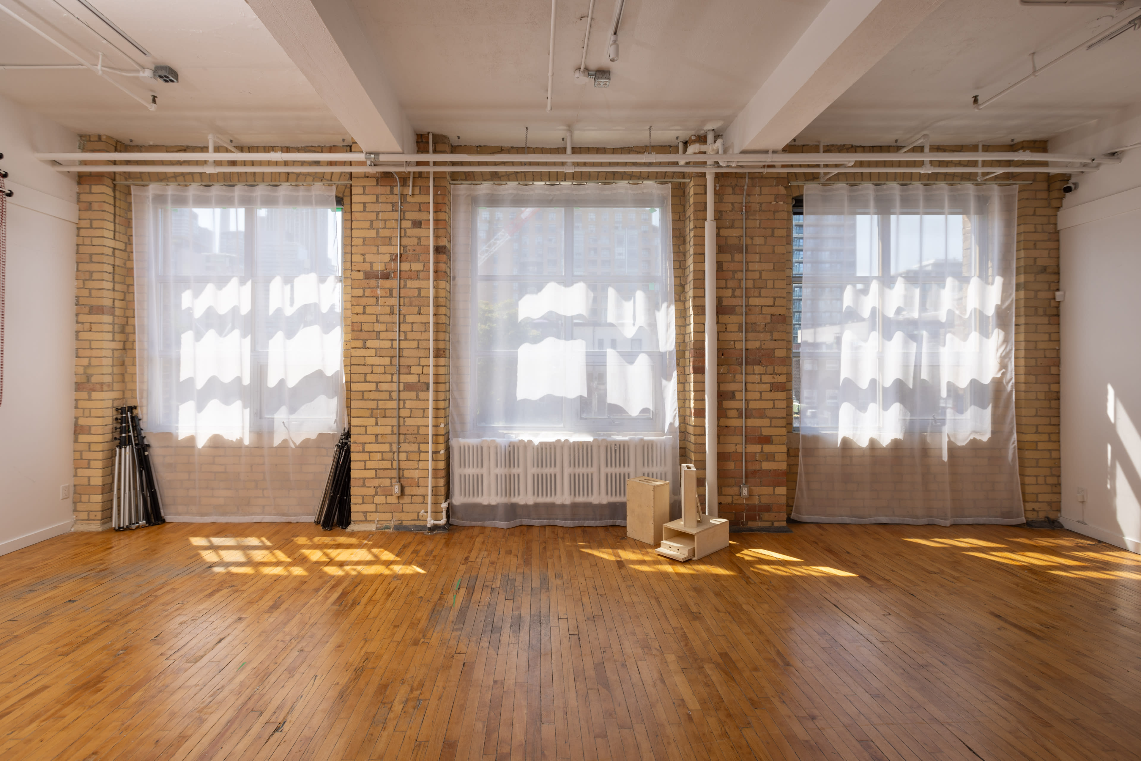 The image shows a bright, spacious room with large windows draped in sheer curtains, revealing wooden floors and brick walls.