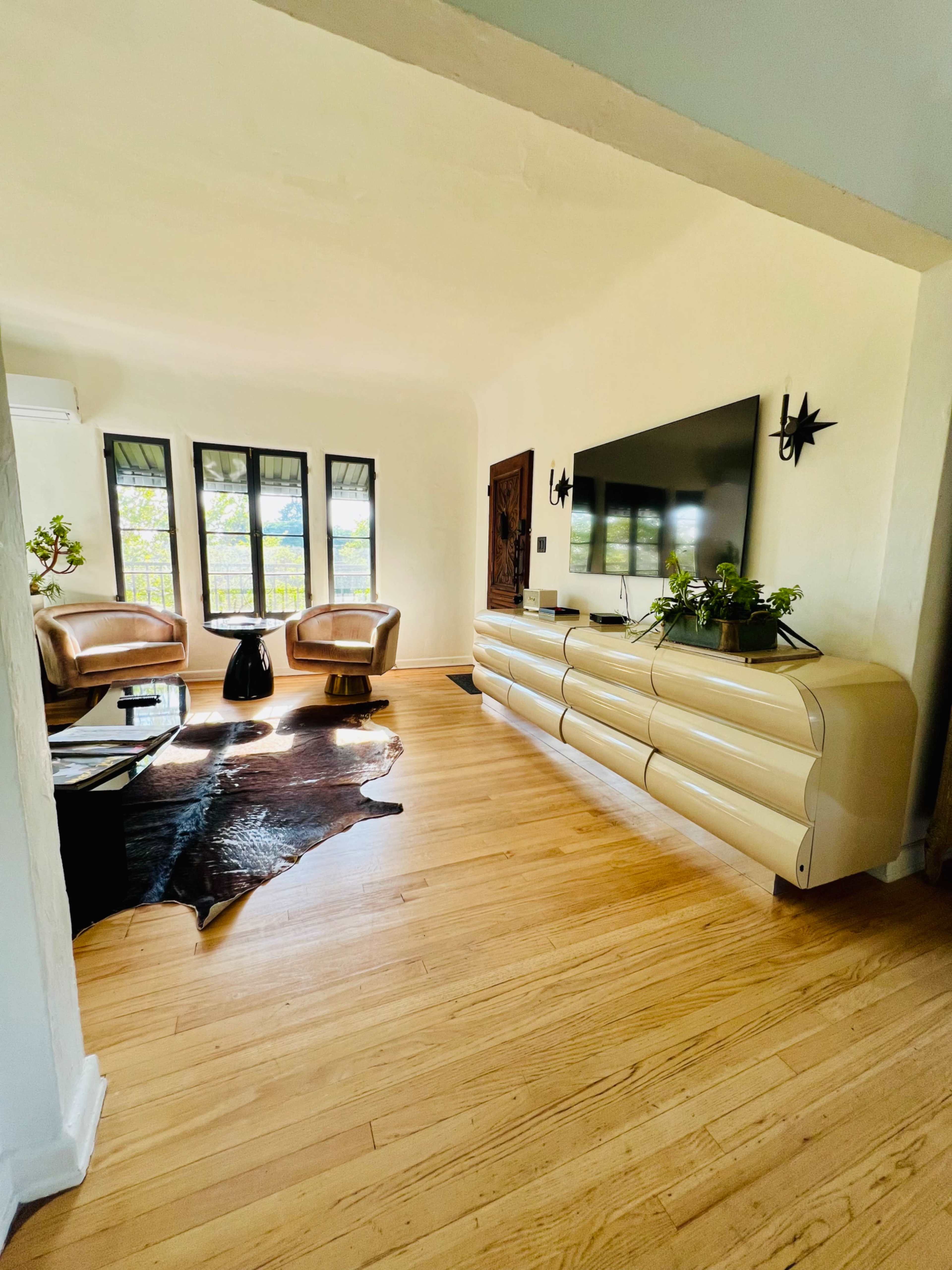 The image shows a modern living room with two brown leather chairs, a large television mounted on a cream-colored cabinet, a black table, and windows providing natural light.