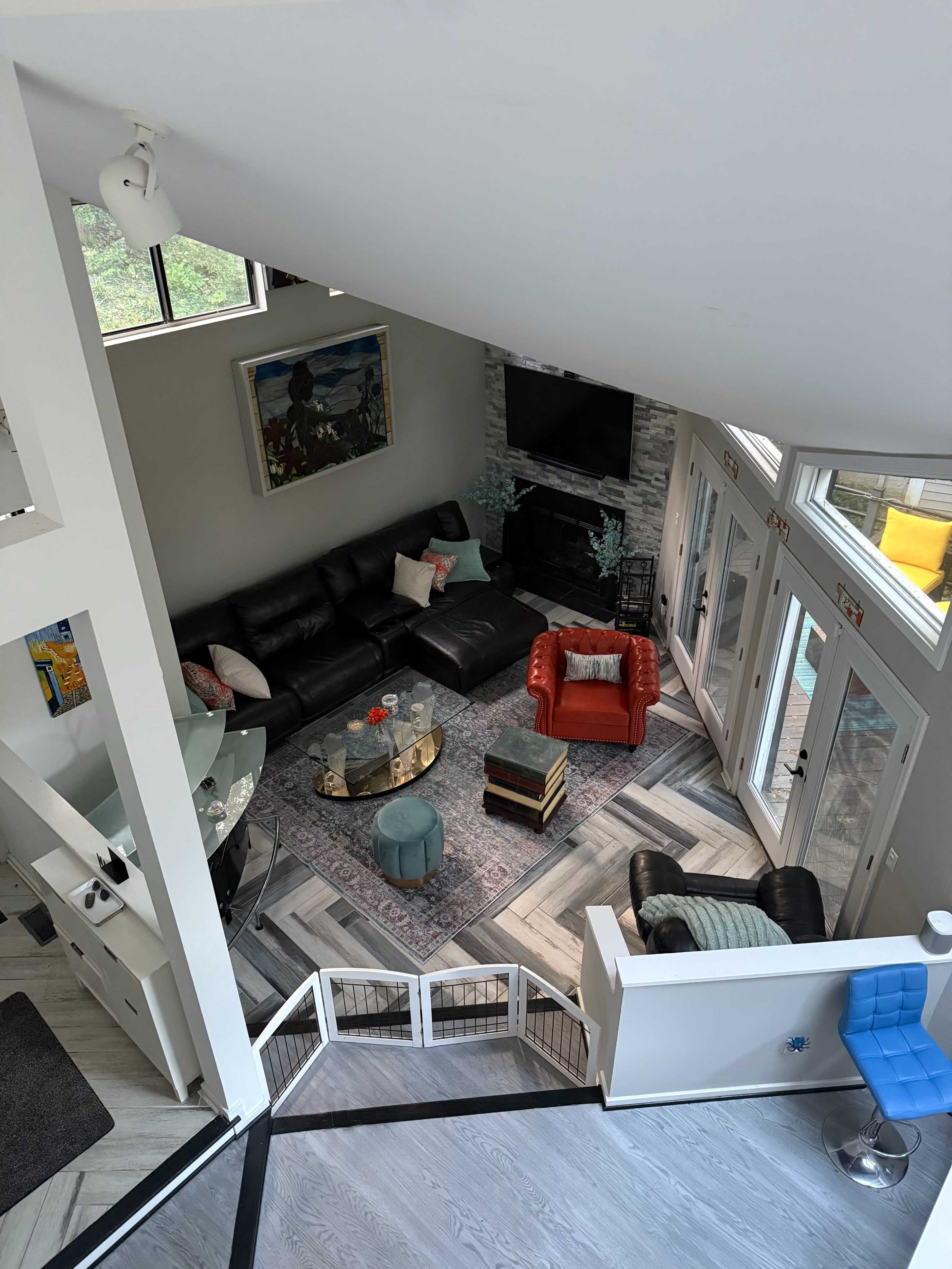 The image shows a modern living room with a black leather sofa, an orange armchair, a round coffee table, and a large window allowing natural light to enter.