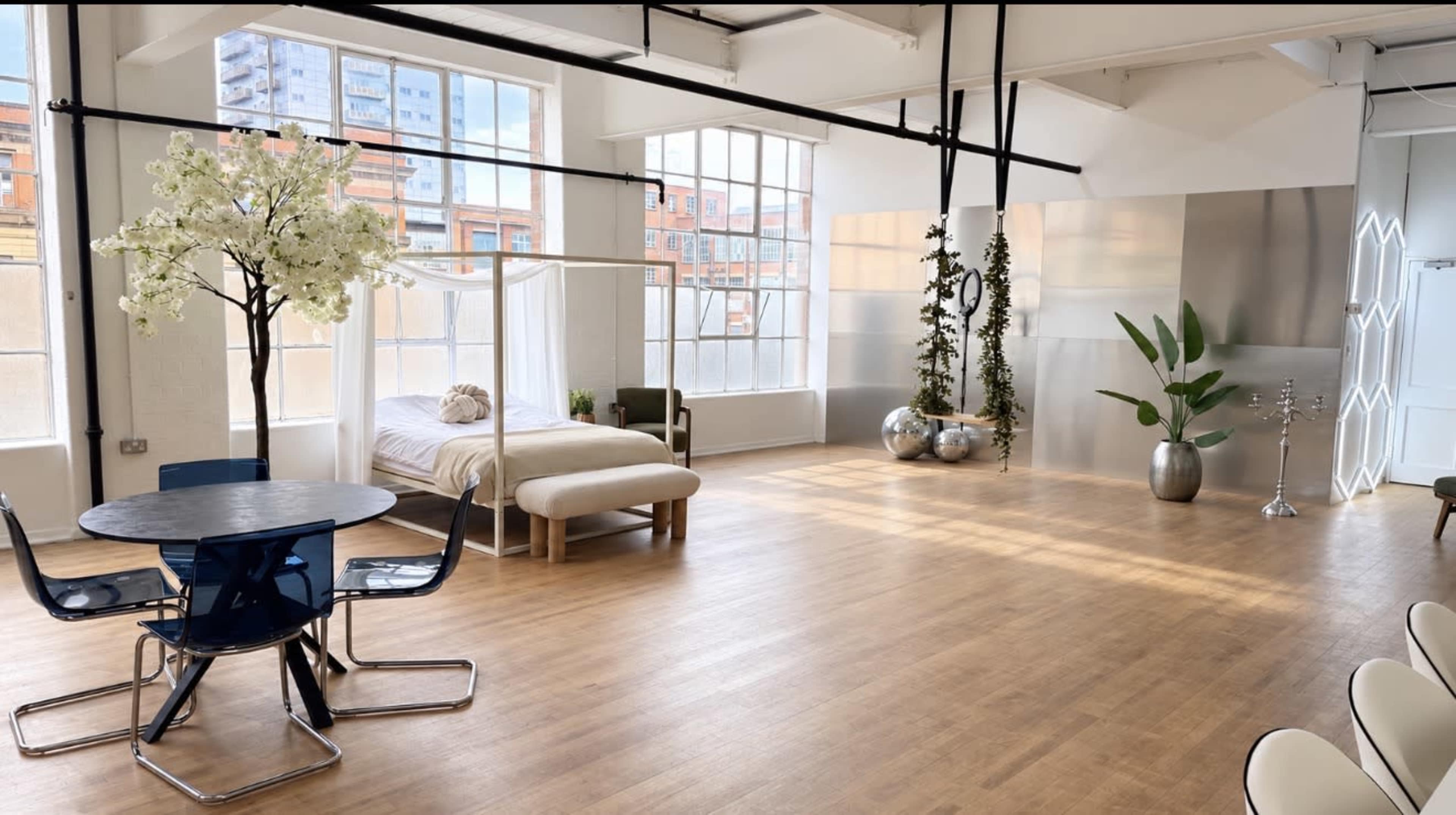 The image shows a spacious, well-lit room featuring a bed with a canopy, a circular dining table, and decorative plants.
