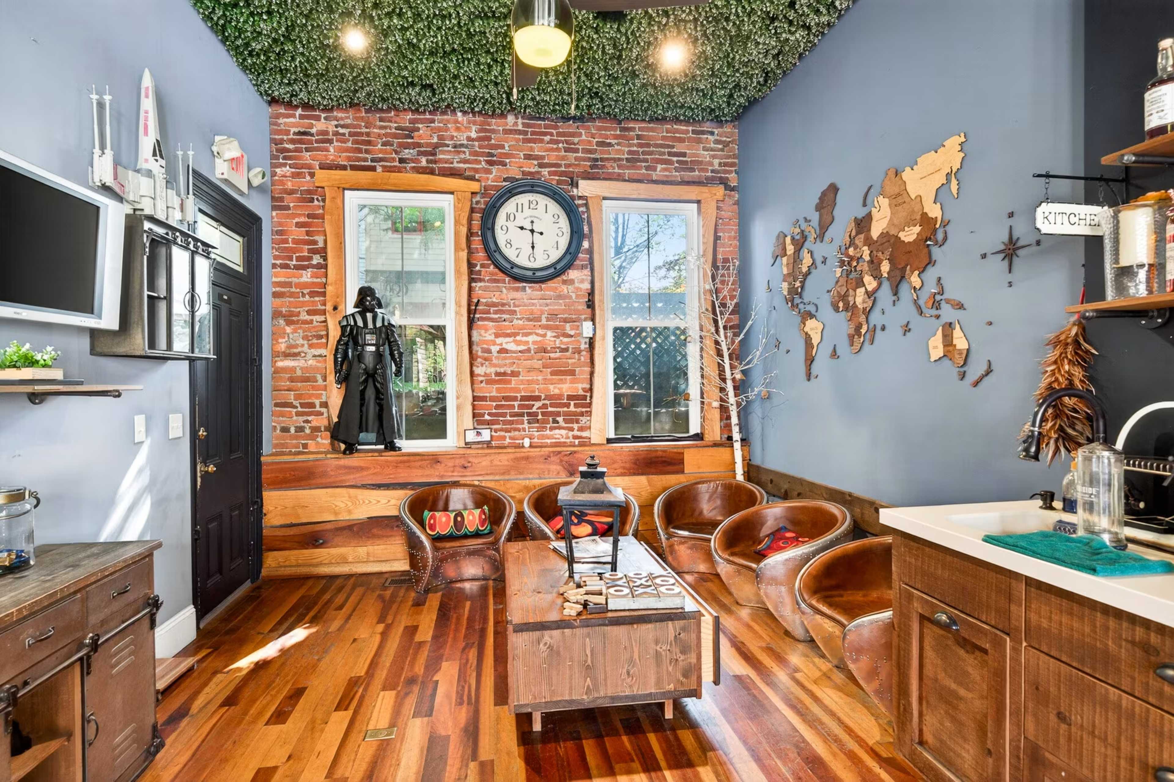 The image shows a room with exposed brick walls, wooden flooring, and a large wall clock, featuring a mix of modern and rustic decor, including a figure resembling a character in dark attire.