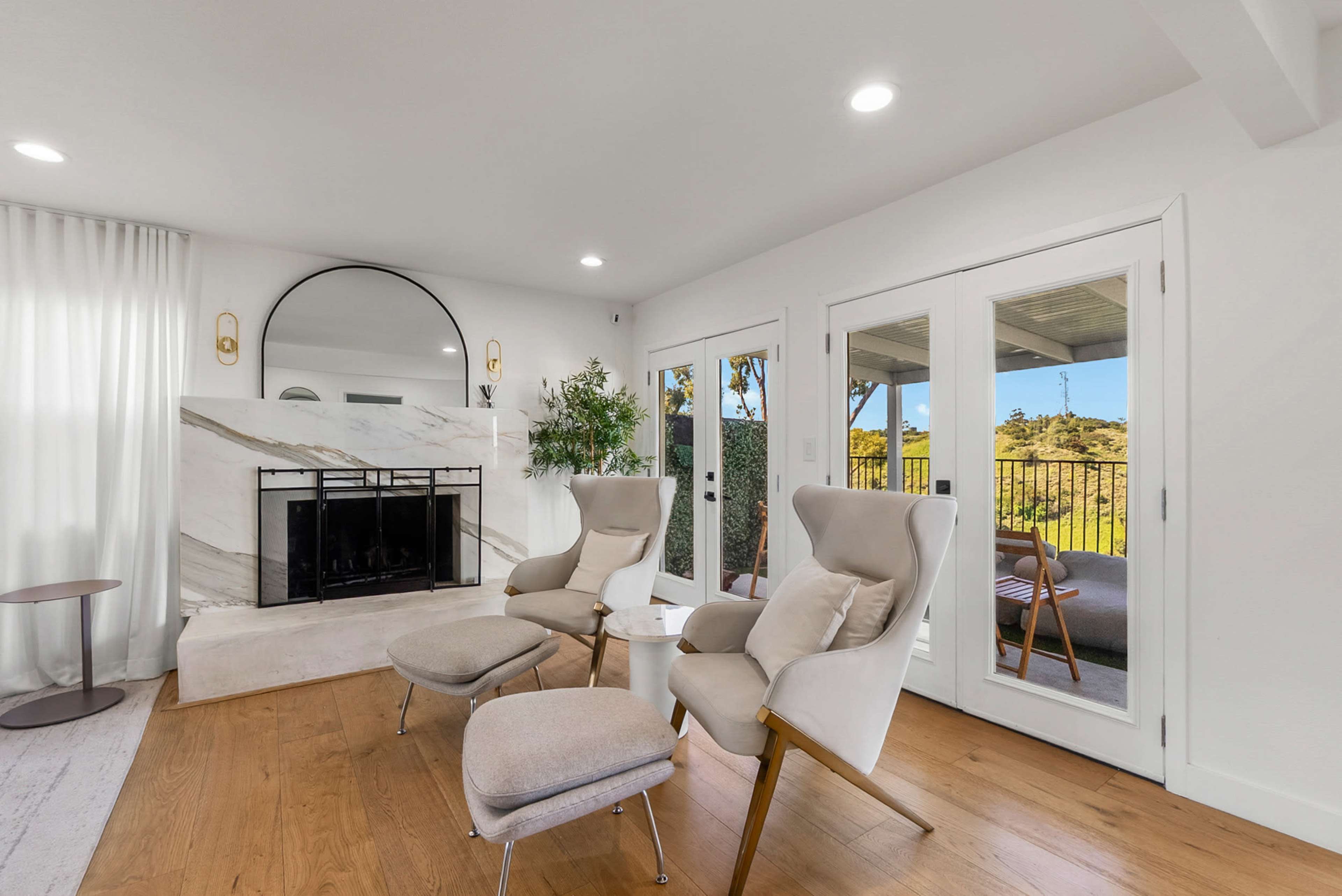 A modern living room features two upholstered chairs facing a marble fireplace, with large glass doors opening to a balcony.