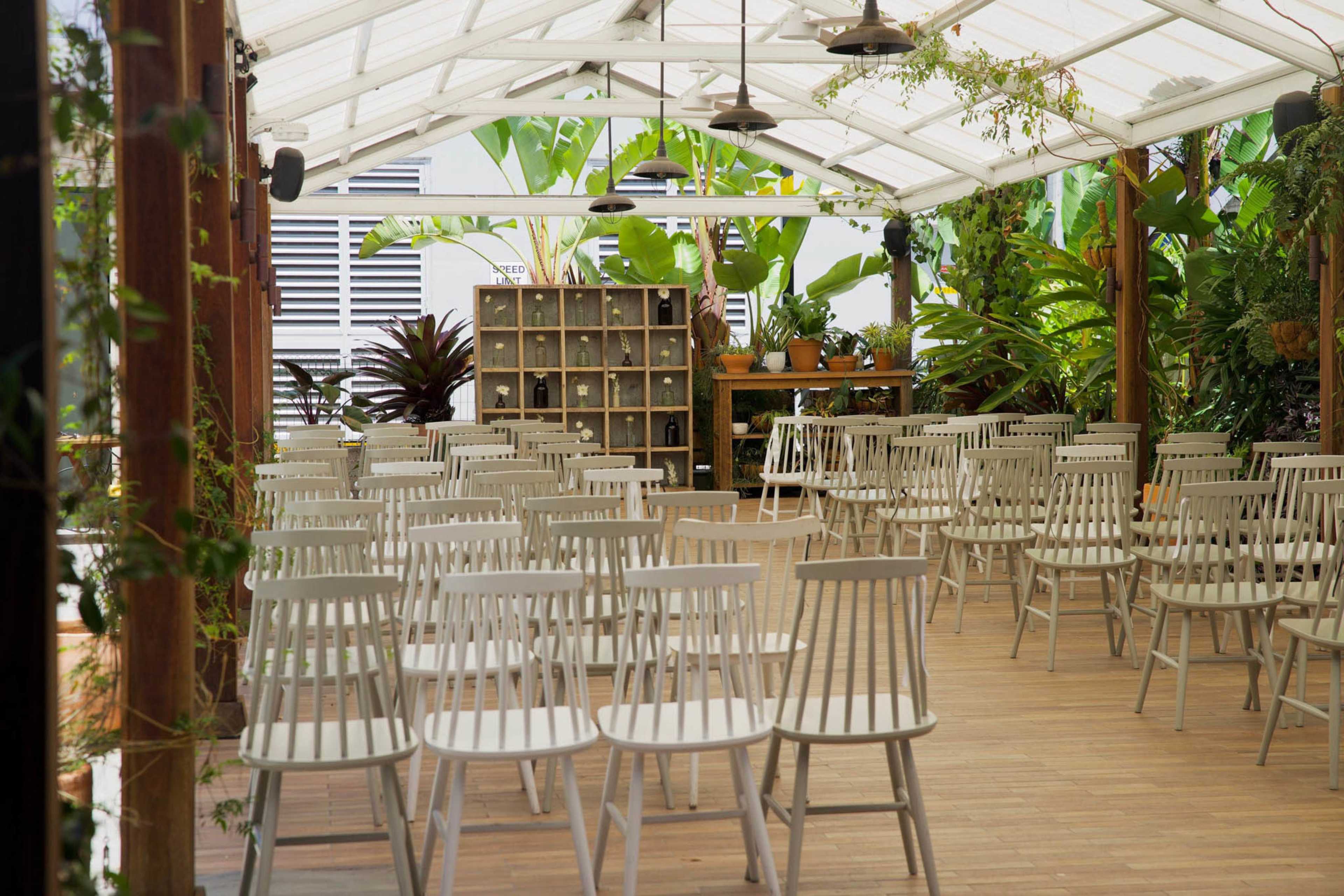 A spacious, airy venue features rows of white chairs arranged for an event, surrounded by greenery and a wooden backdrop.