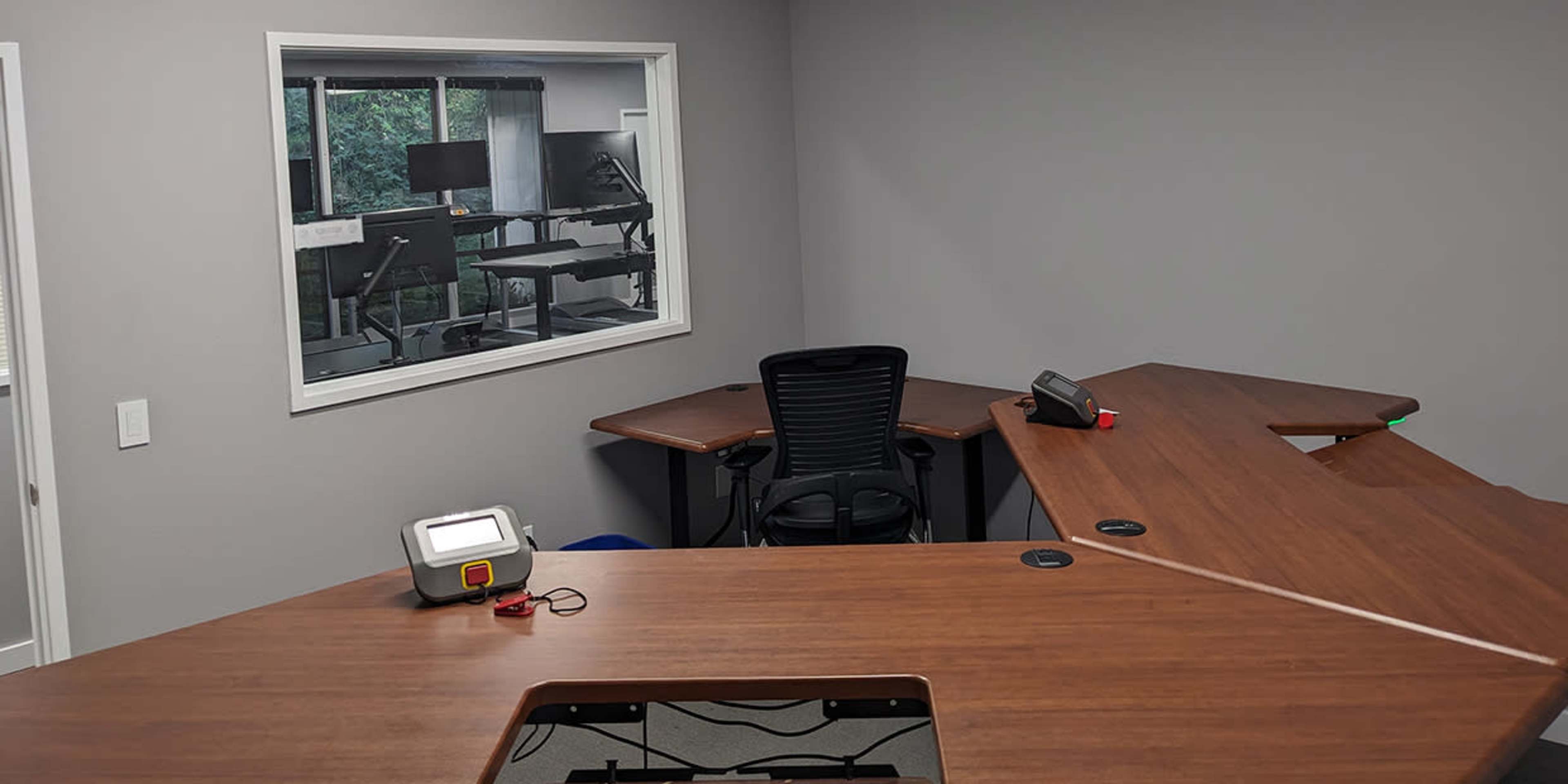 A modern office space with two wooden desks positioned at an angle and a large window overlooking another room with office furniture.