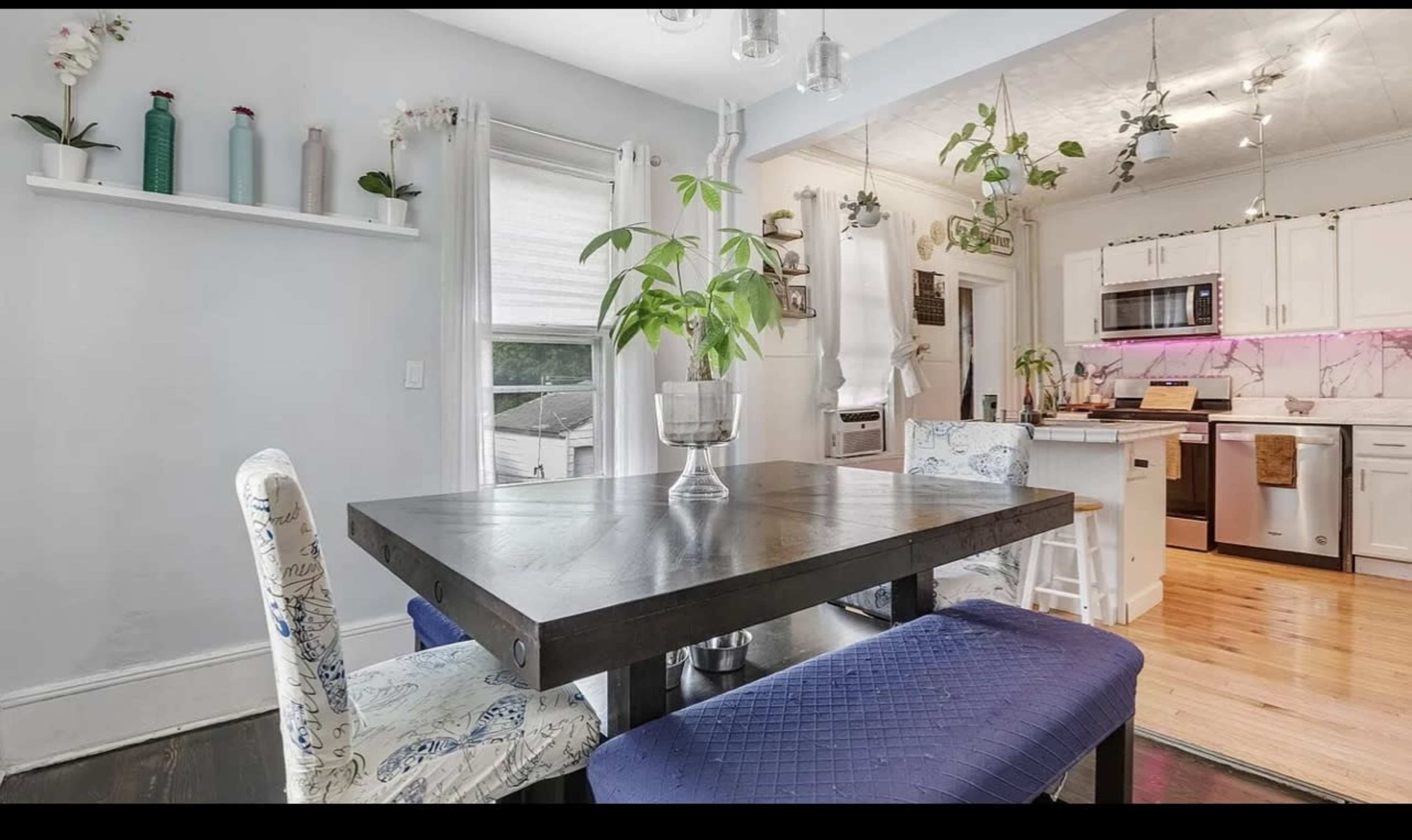 A dining area with a dark table and two upholstered chairs, adjacent to a kitchen featuring modern appliances and decorative plants.