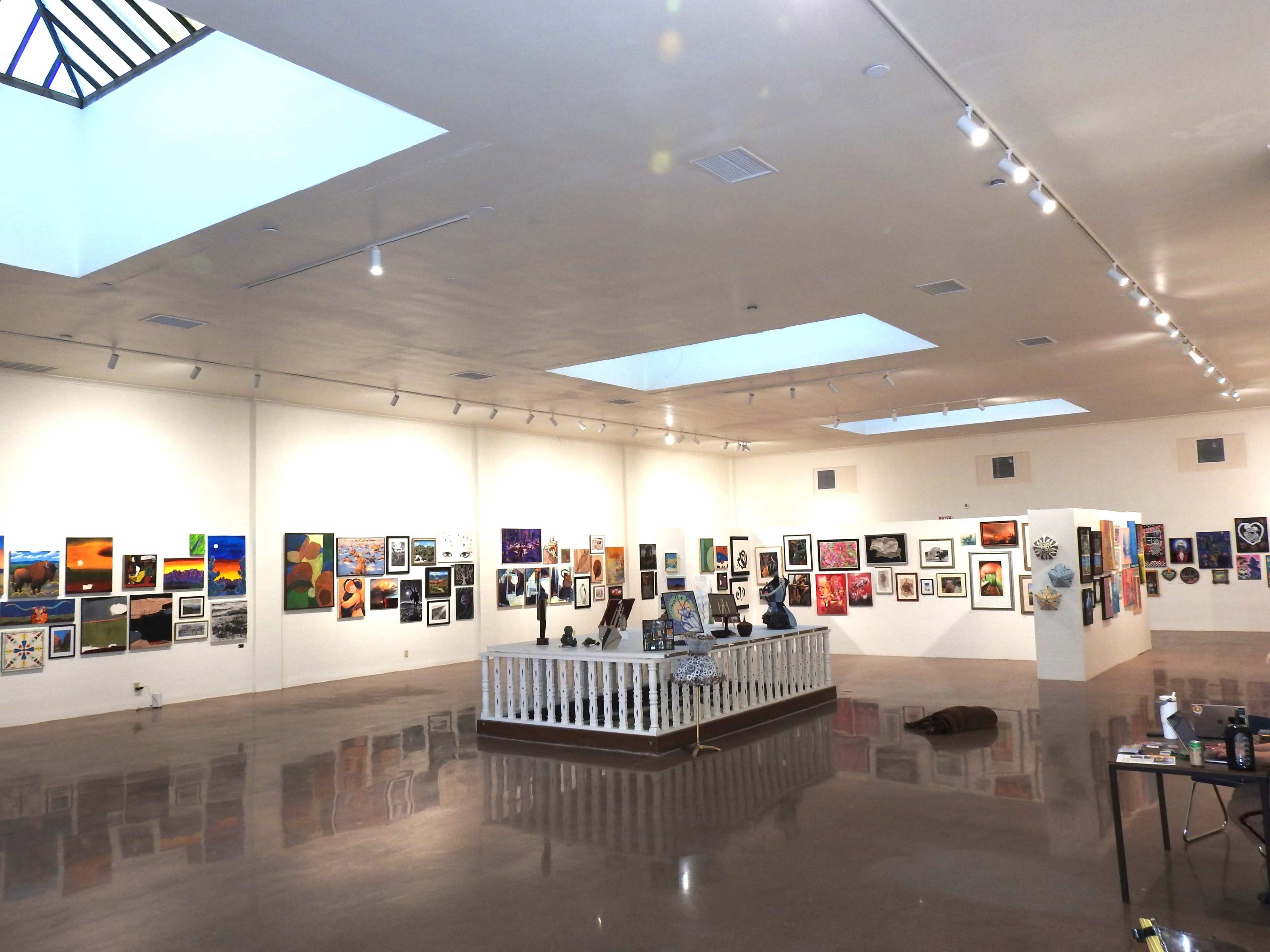 The image shows an art gallery with various paintings displayed on the walls and a central installation area featuring sculptures.