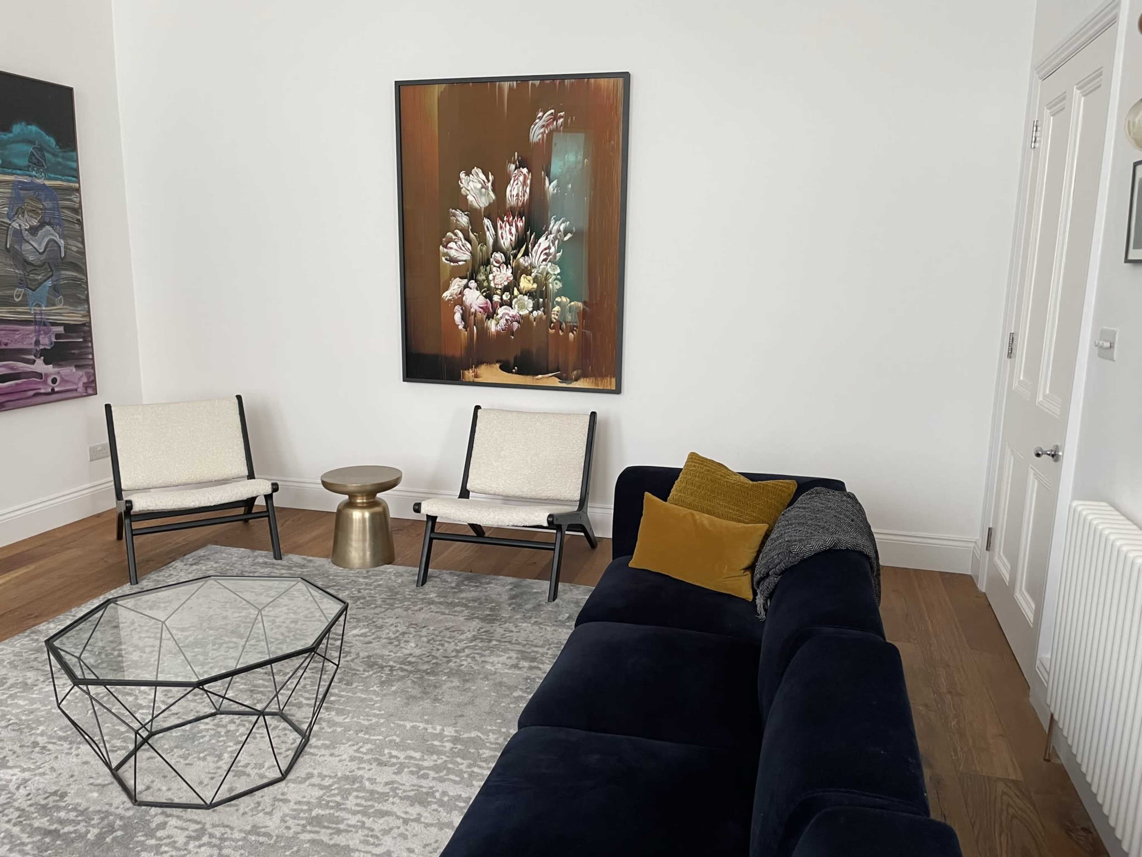 The image shows a modern living room with a dark sofa, two light-colored chairs, a glass coffee table, and a large floral artwork on the wall.
