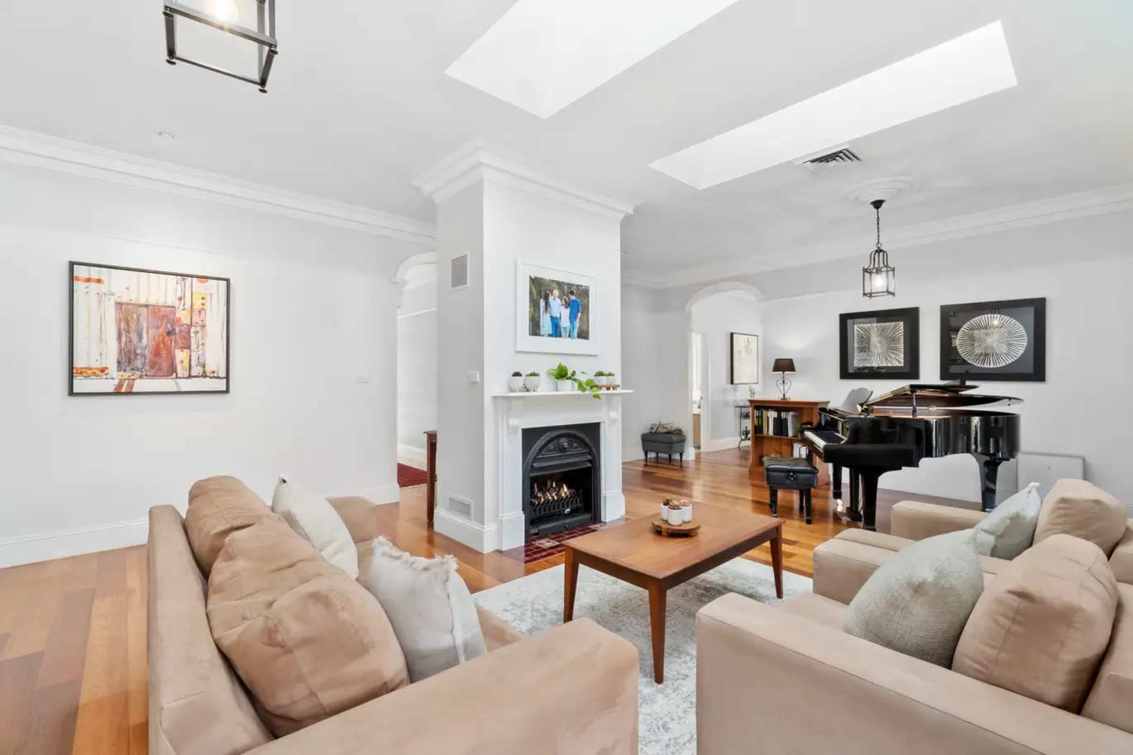 The image shows a bright living room with a fireplace, two beige sofas, a coffee table, and a grand piano in the corner.
