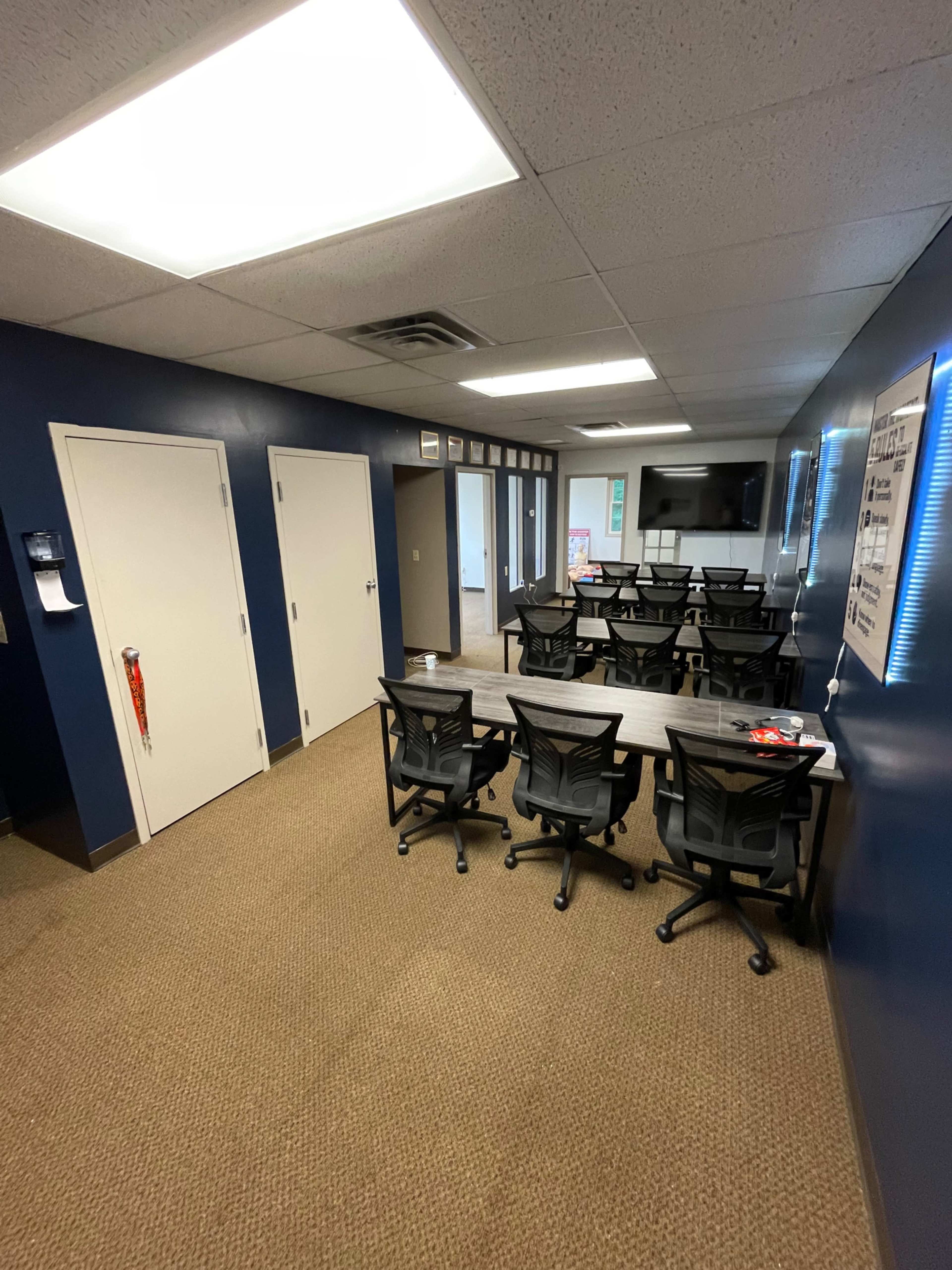 The image shows a classroom or meeting room with multiple tables and chairs arranged, beige carpet flooring, and dark blue walls.