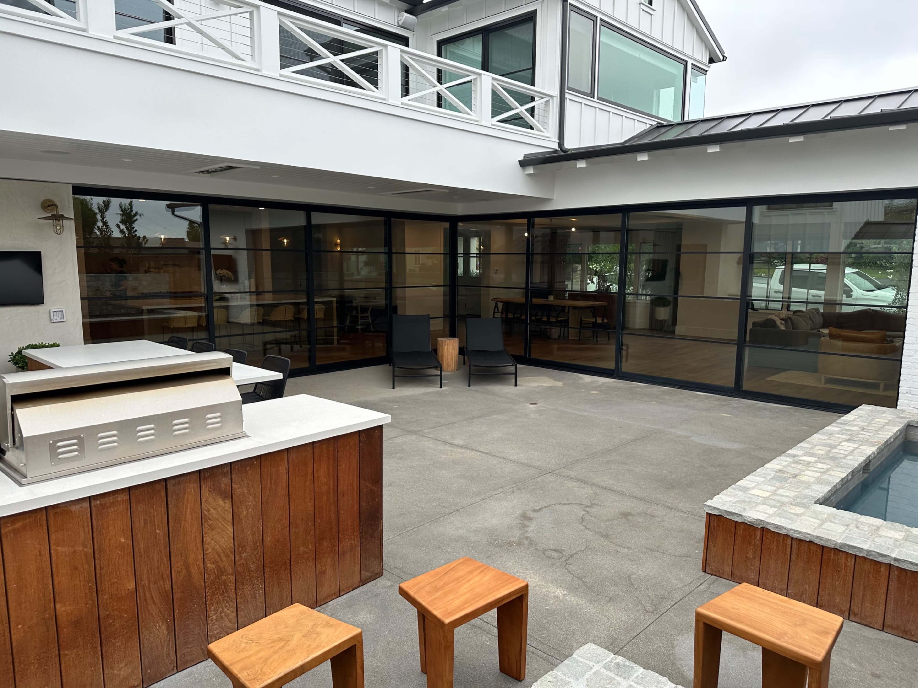 The image shows a modern outdoor space featuring a wooden bar area, a pool, and several wooden stools adjacent to a house with large glass sliding doors.