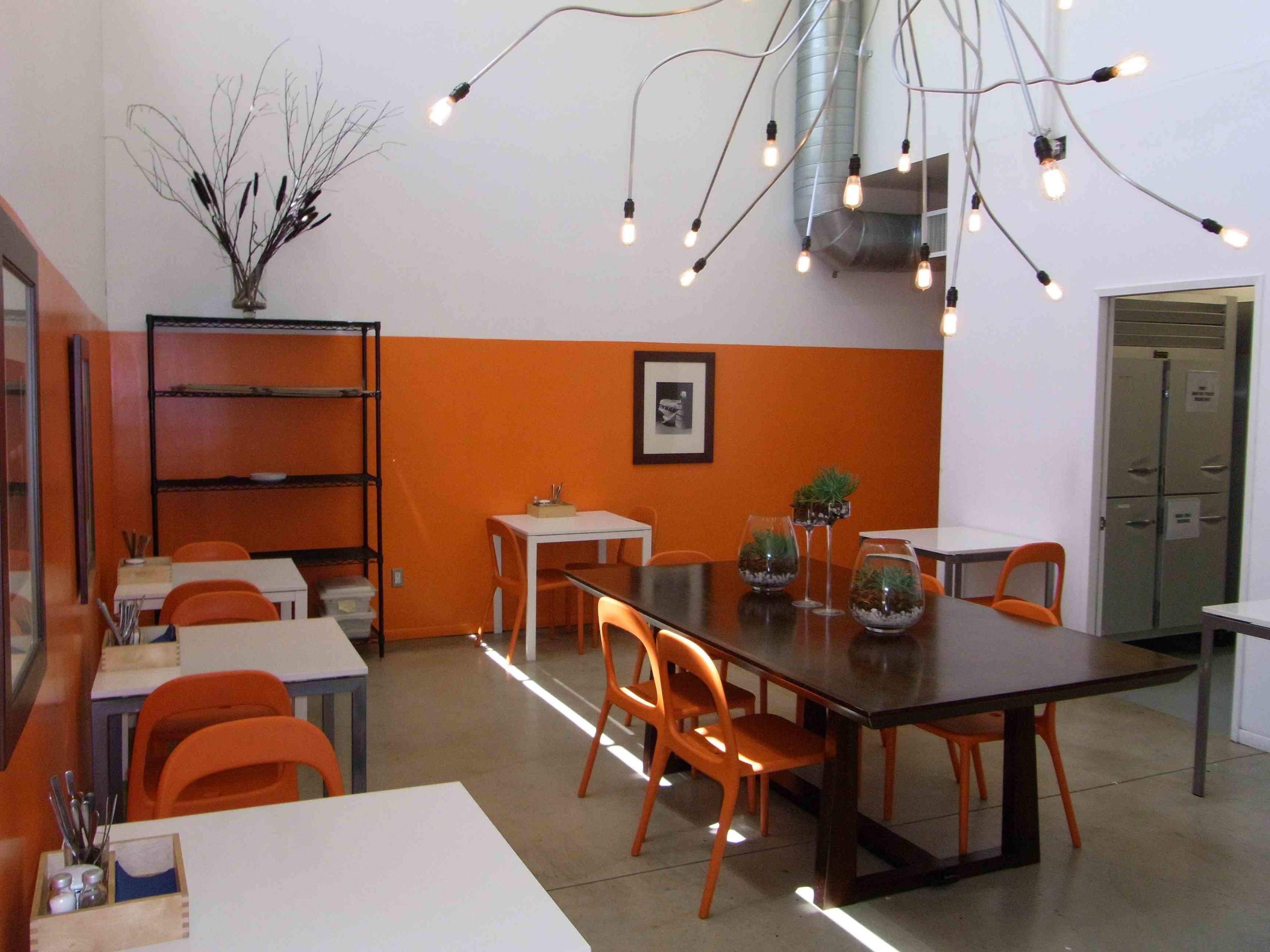A modern workspace featuring orange accent walls, a large wooden table, and several white tables and chairs, all under unique pendant lighting.
