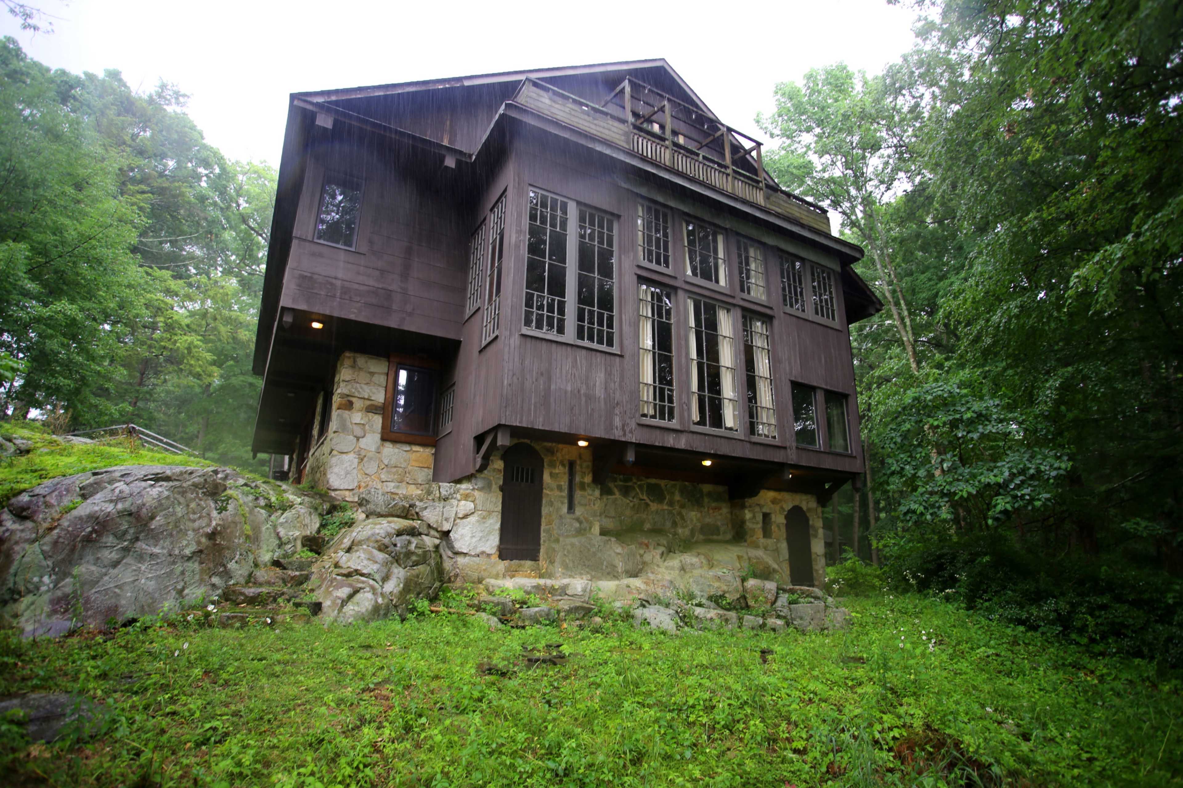 A three-story wooden house sits on a rocky foundation surrounded by dense greenery.