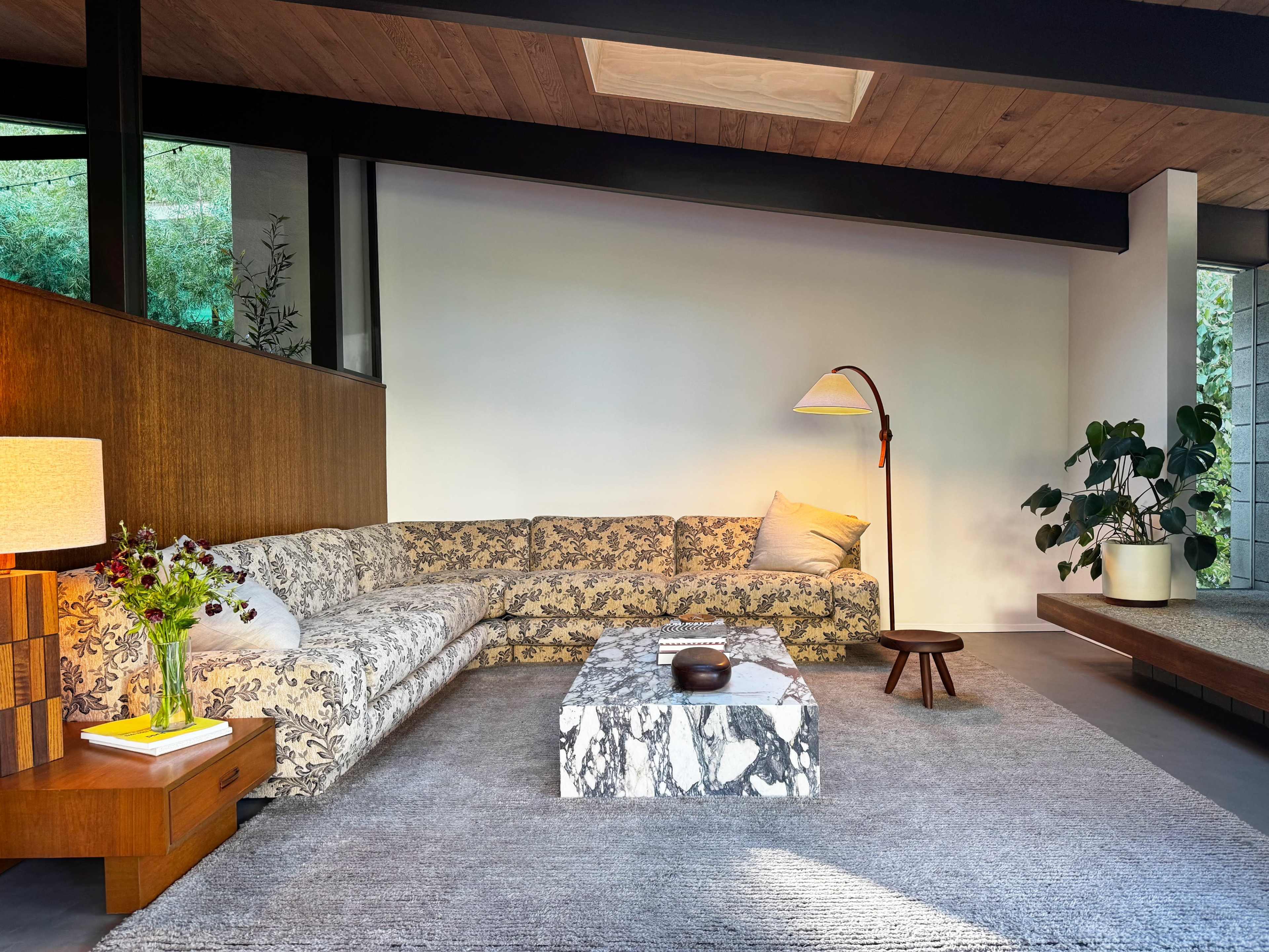 The image depicts a modern living room featuring a floral-patterned sectional sofa, a marble coffee table, a wooden side table, and a floor lamp, surrounded by greenery and natural light.