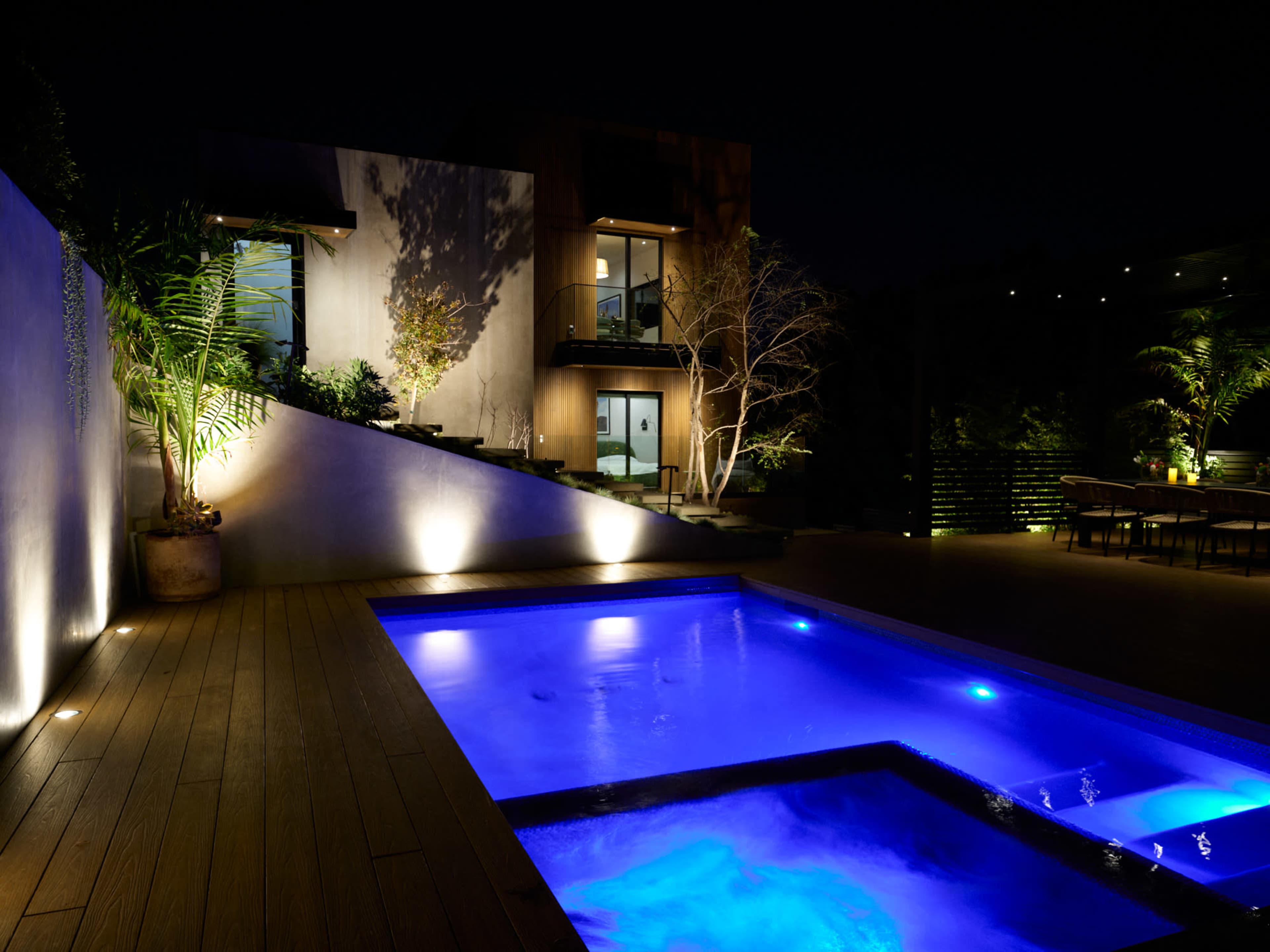 A modern backyard with a illuminated pool and a multi-level home lit by exterior lighting at night.