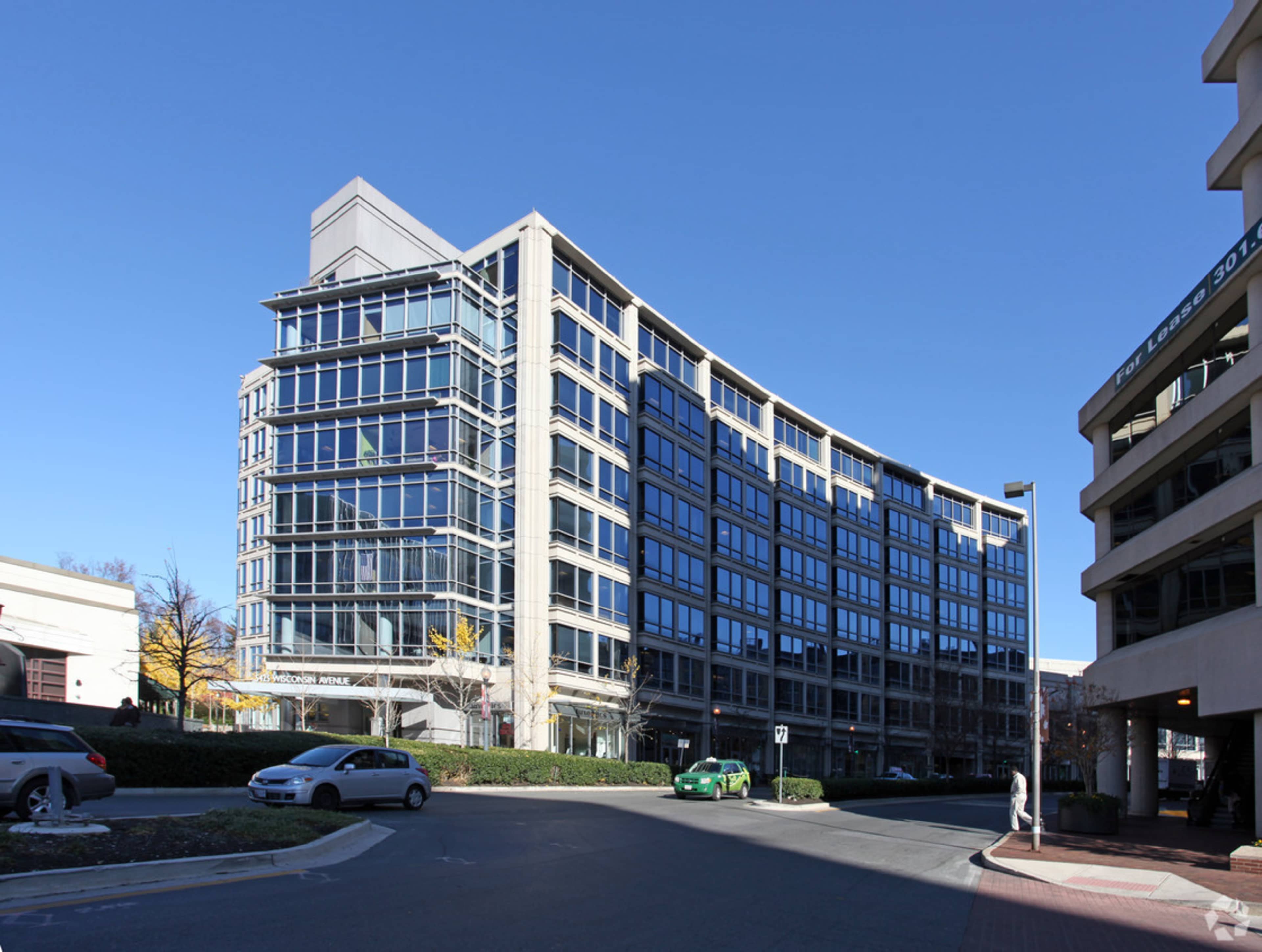 The image shows a modern, multi-story office building with large glass windows and a curved structure, located at the end of a street with parked cars.