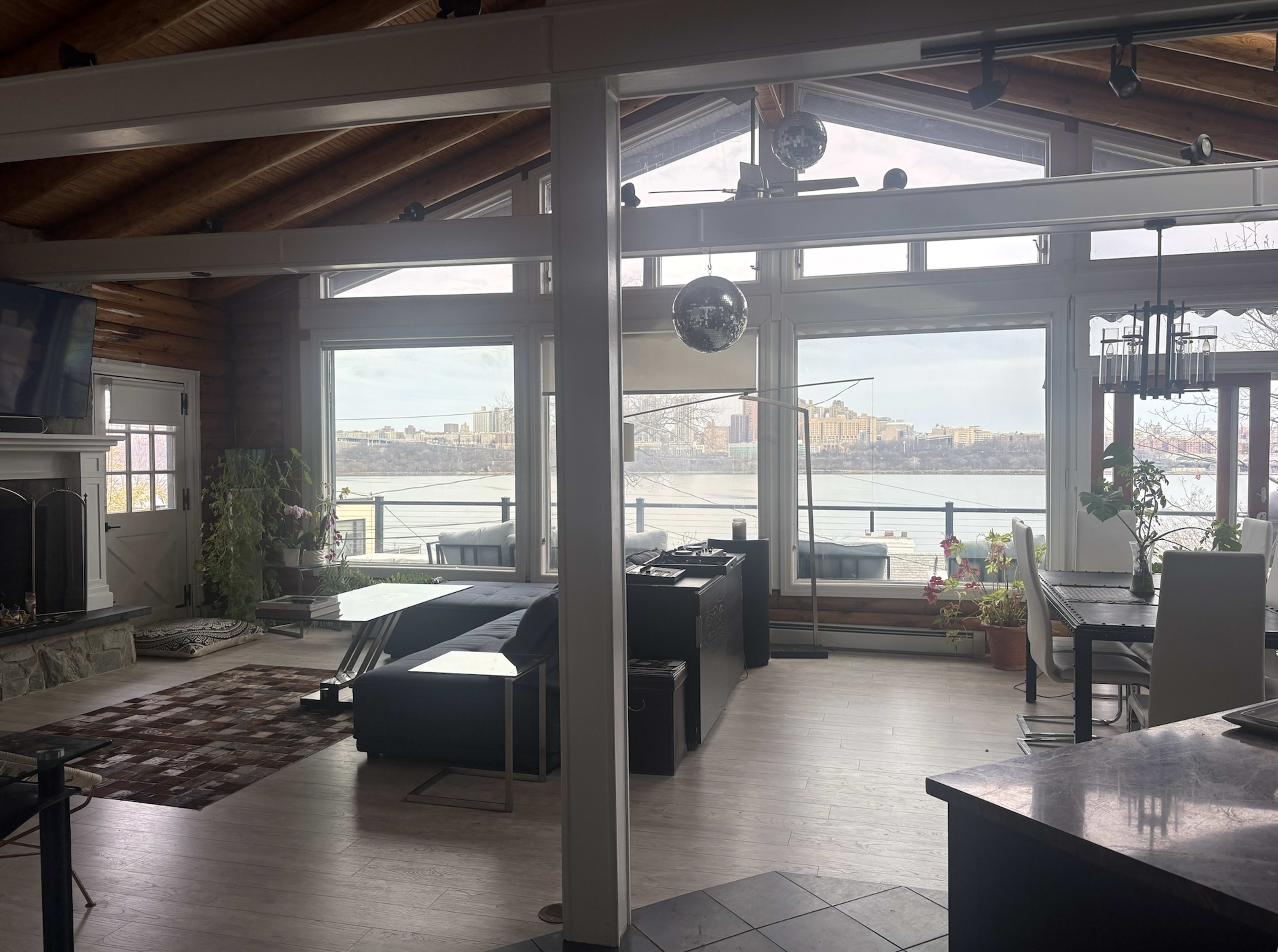 The image shows a spacious living area with large windows overlooking a body of water and a city skyline, featuring modern furnishings and wooden beams.