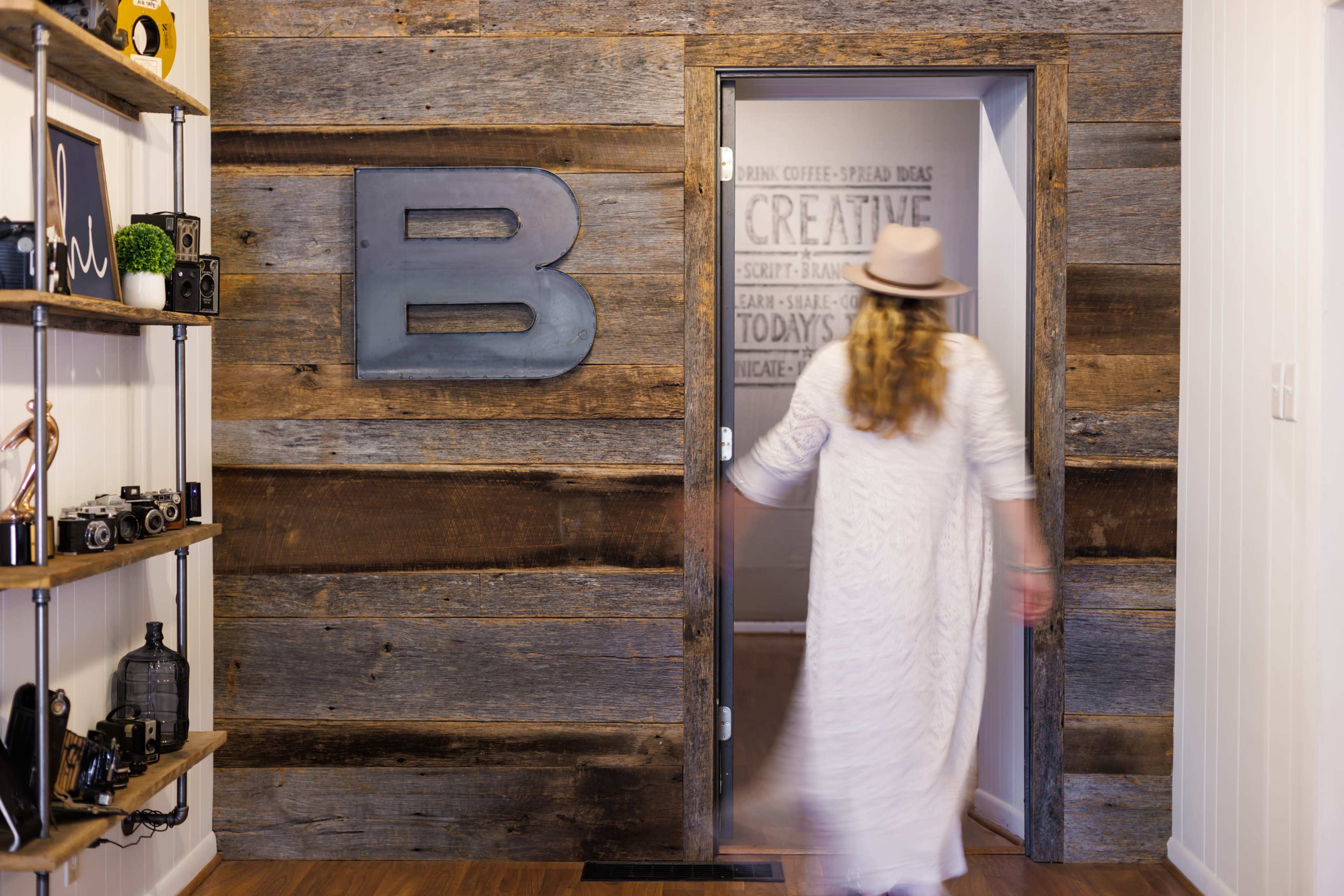A person in a long white dress walks towards a partially open door with a metallic letter "B" on a rustic wooden wall.