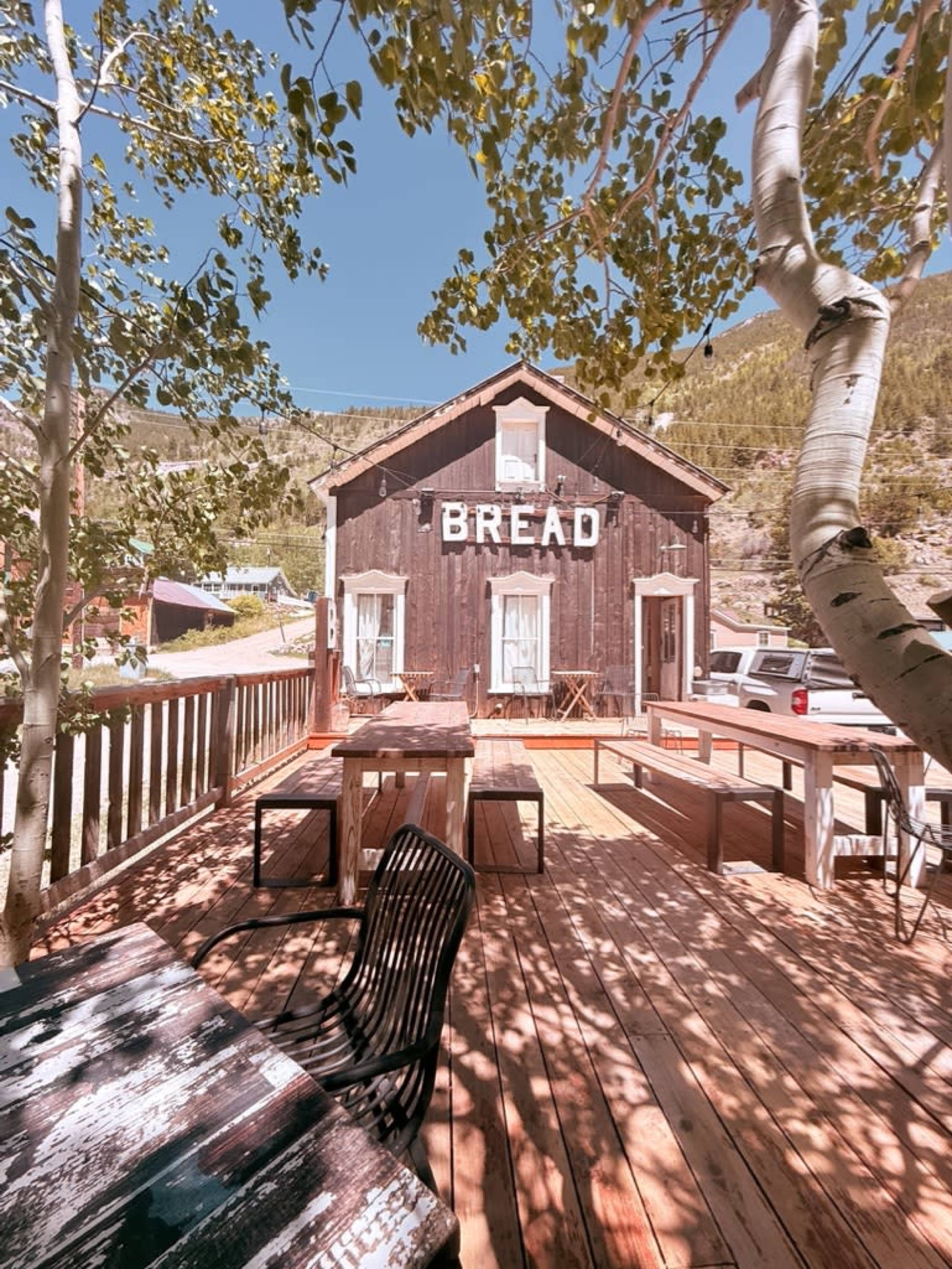 A rustic wooden building with the word "BREAD" displayed prominently on its facade is surrounded by green trees and a spacious wooden deck with outdoor seating.