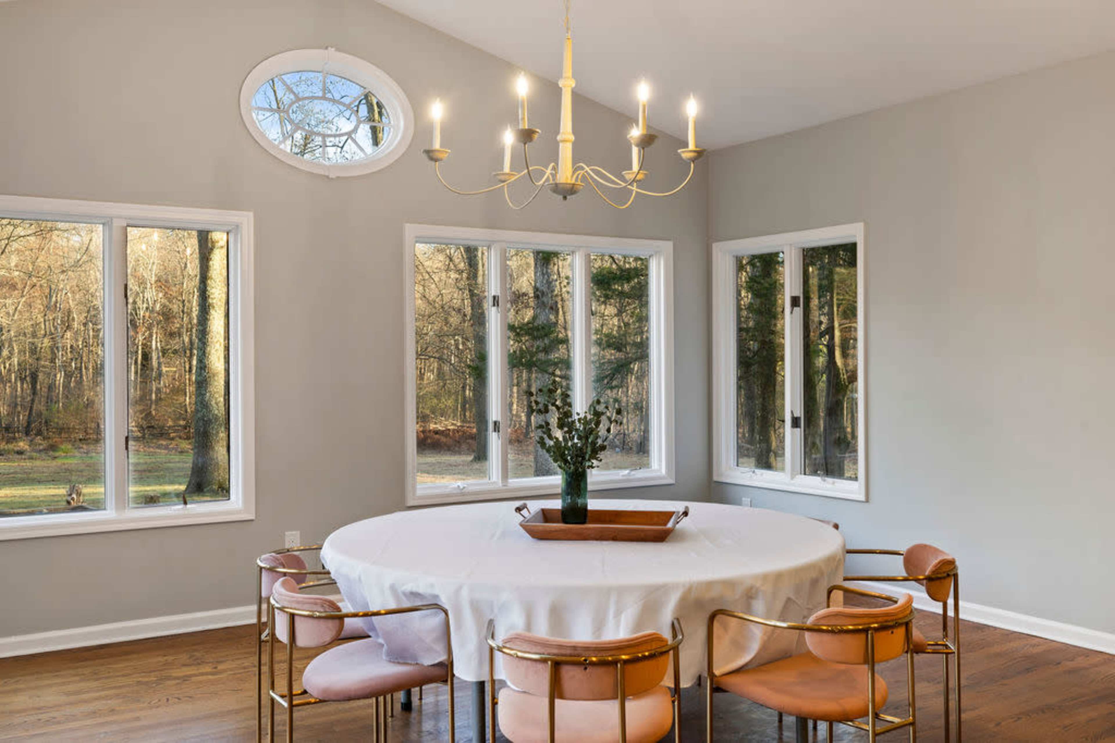 A round dining table with a white tablecloth is surrounded by six gold-framed chairs in a well-lit room featuring large windows that overlook a wooded area.