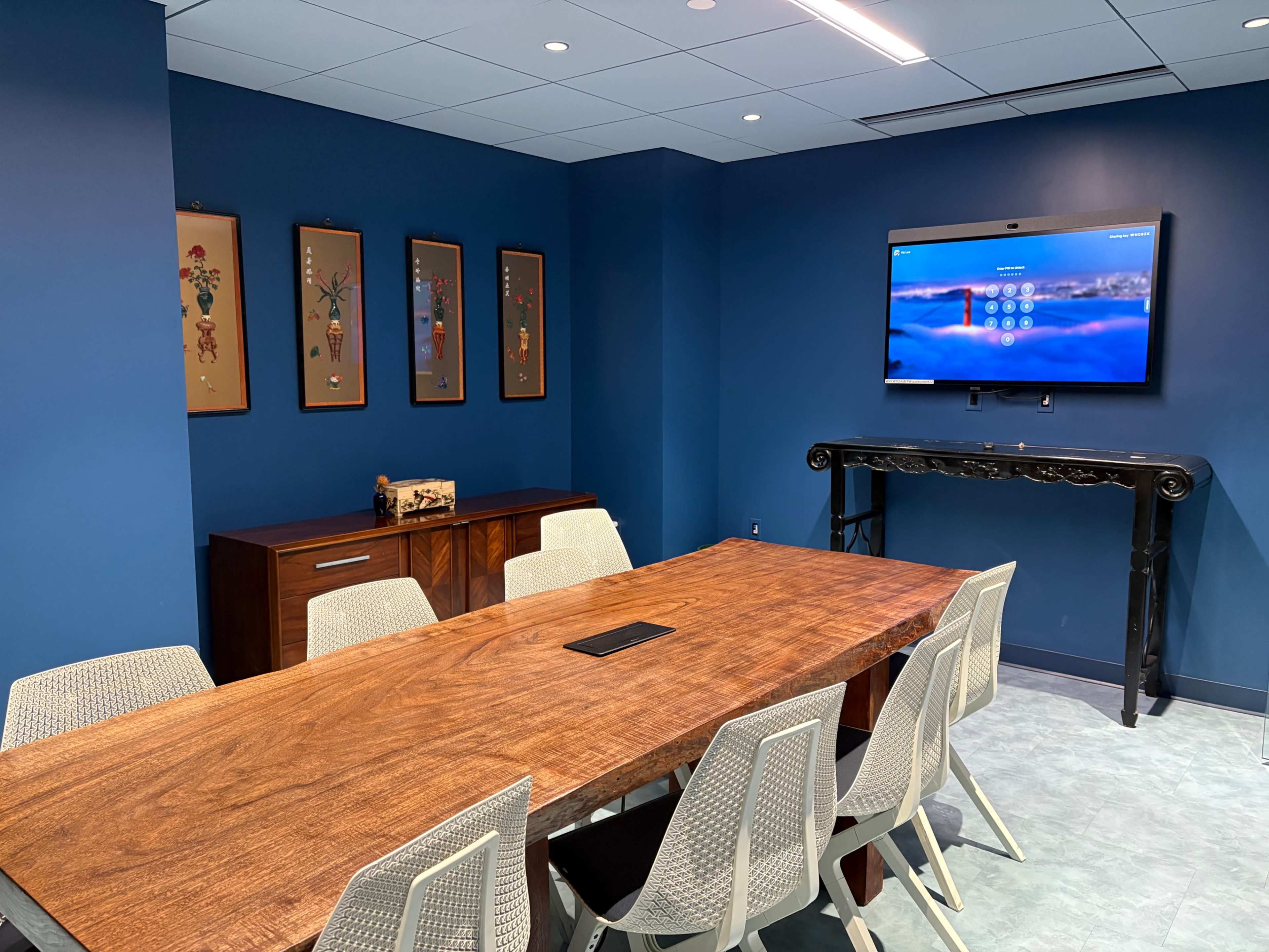 A modern conference room features a wooden table surrounded by white chairs, with artwork on the walls and a screen mounted at the front.