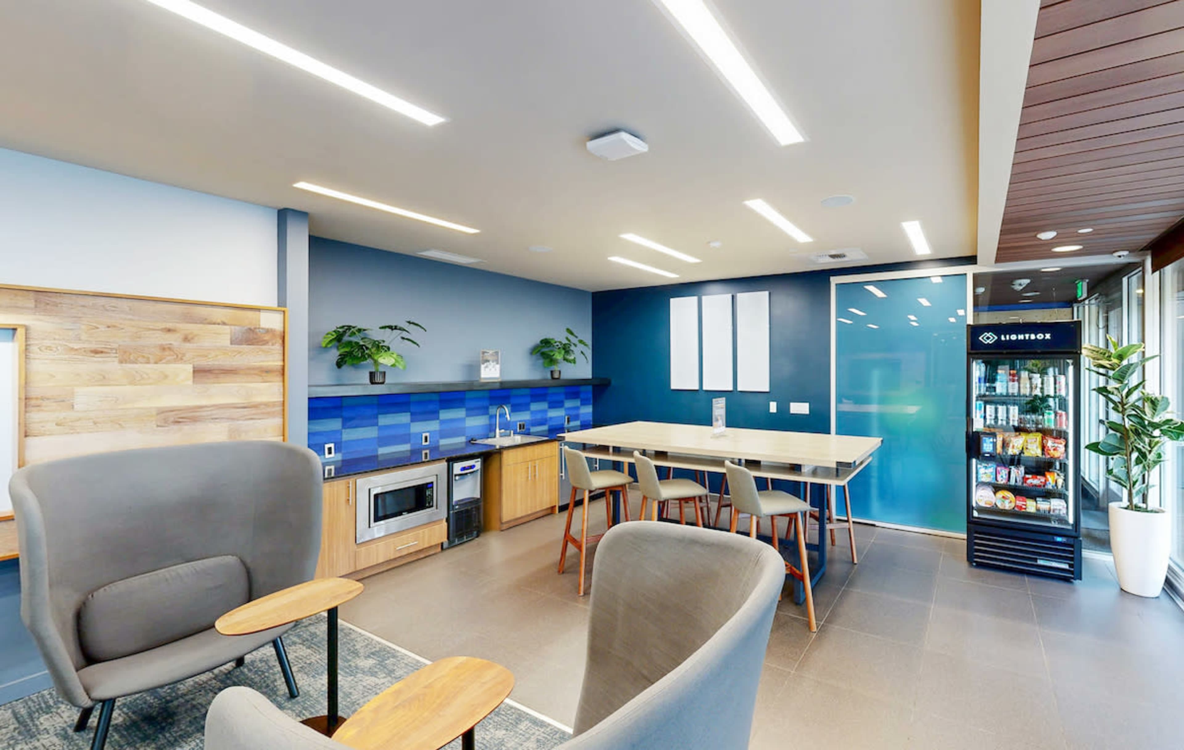 The image shows a modern kitchen area with a blue tiled backsplash, a long wooden table surrounded by chairs, and a beverage refrigerator.