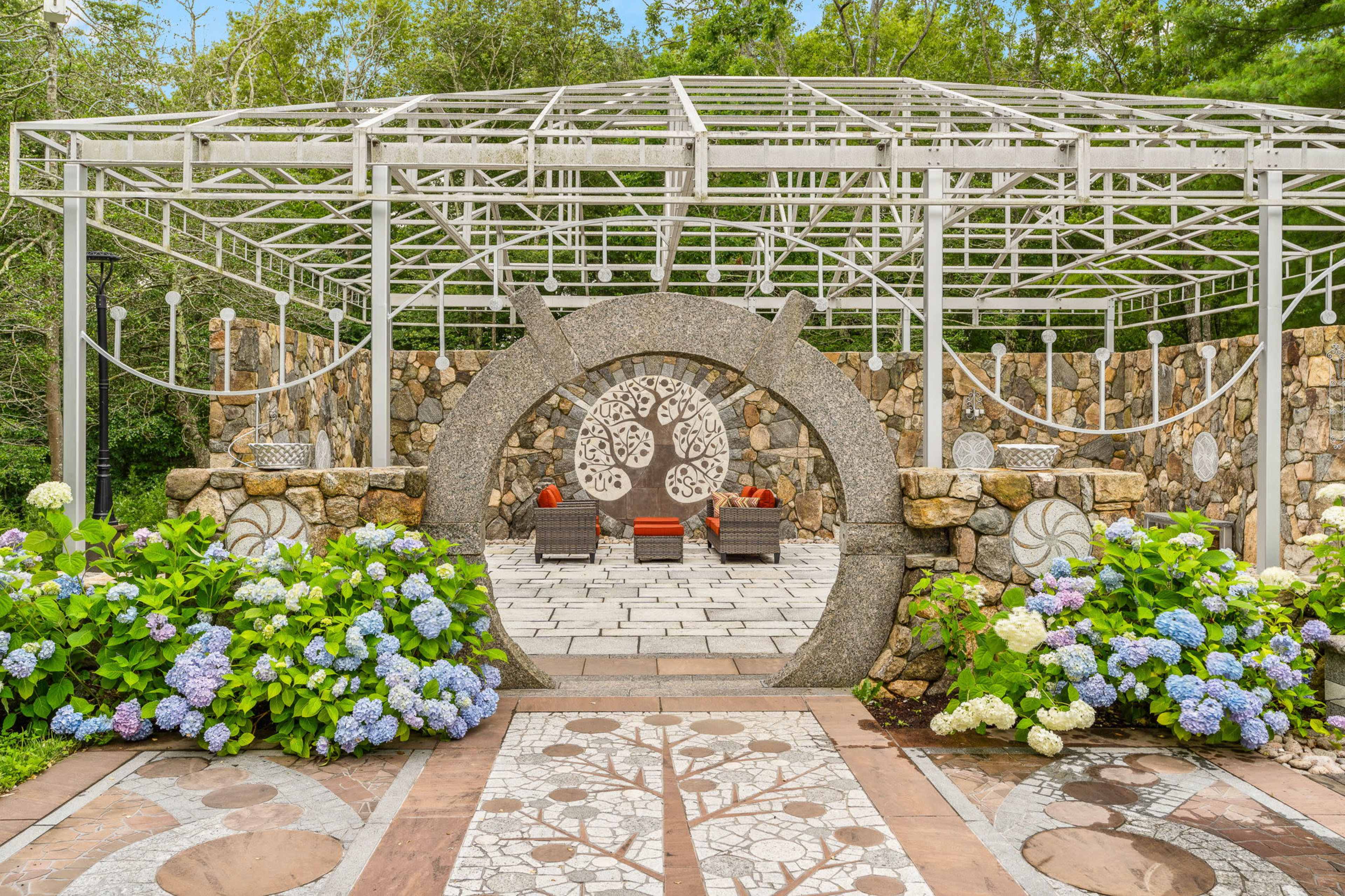 The image features a landscaped garden with a stone pathway, a circular stone archway, and a pergola structure above two seating areas surrounded by hydrangea plants.