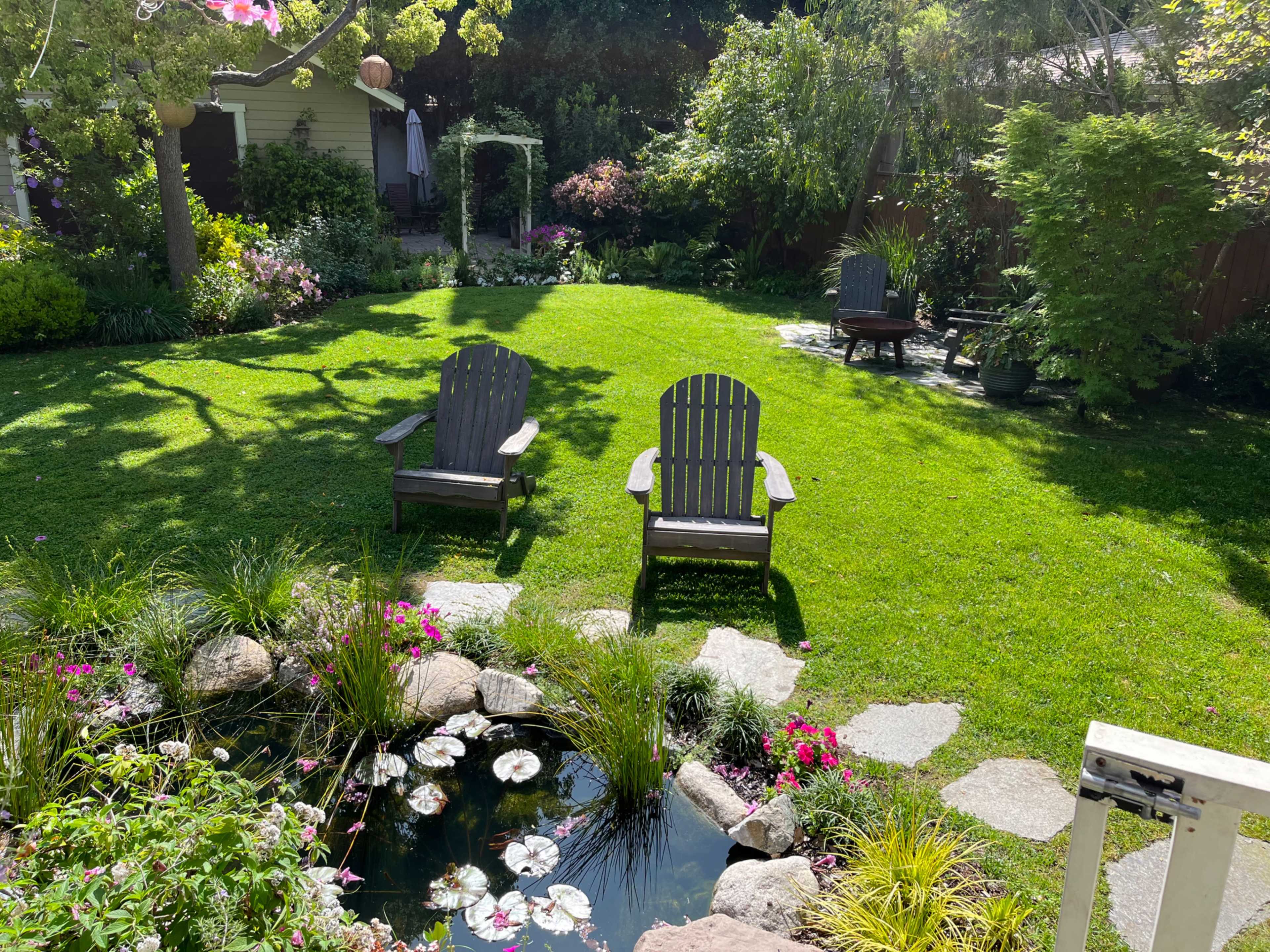 A well-manicured garden with two wooden adirondack chairs facing a small pond surrounded by greenery and flowers.