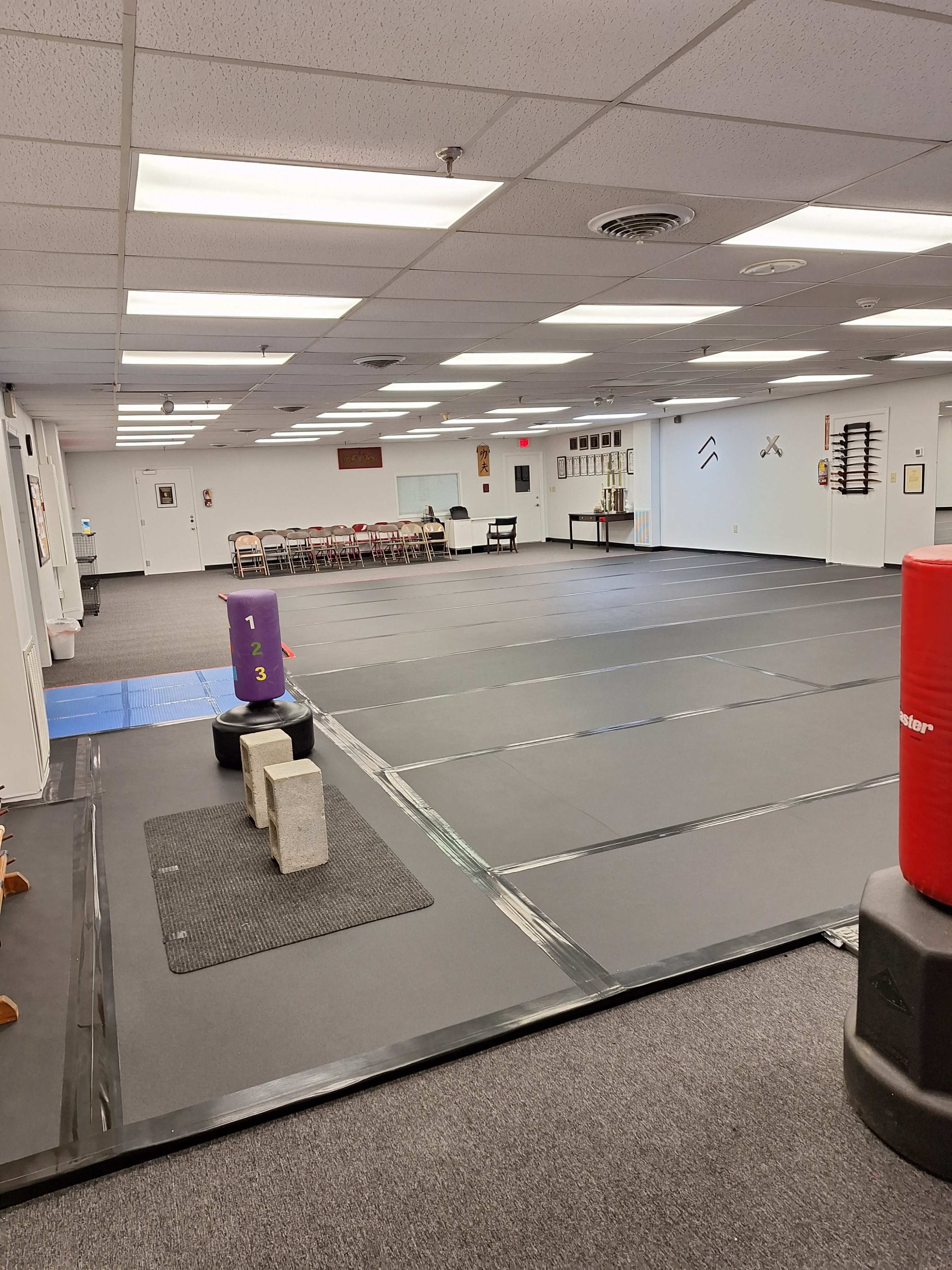 The image shows a large, empty martial arts training hall with padded flooring and various training equipment arranged neatly.