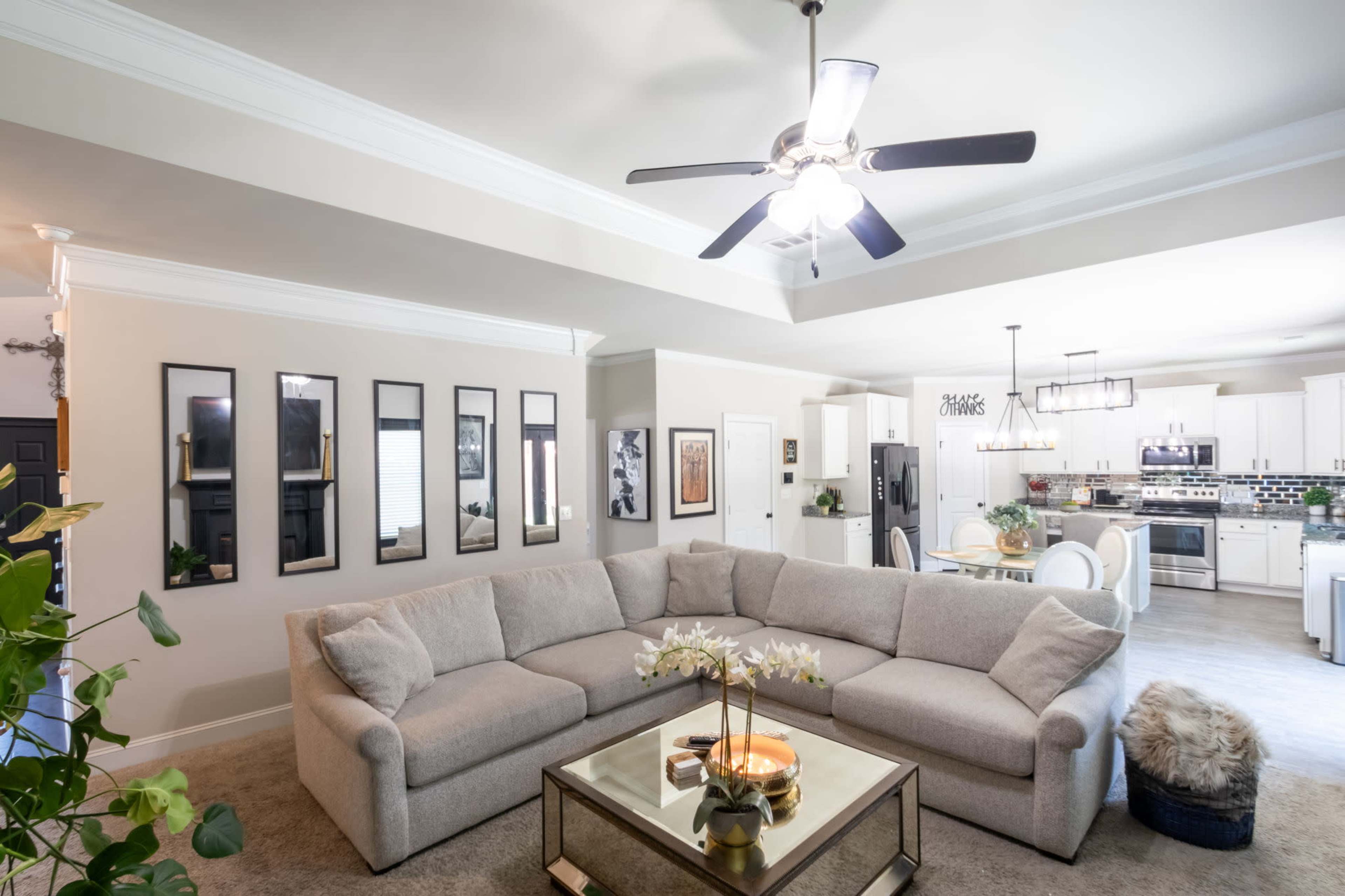 The image shows a modern living room with a large, light-colored sectional sofa, a glass coffee table, and a ceiling fan, open to a kitchen area with white cabinets and green accents.